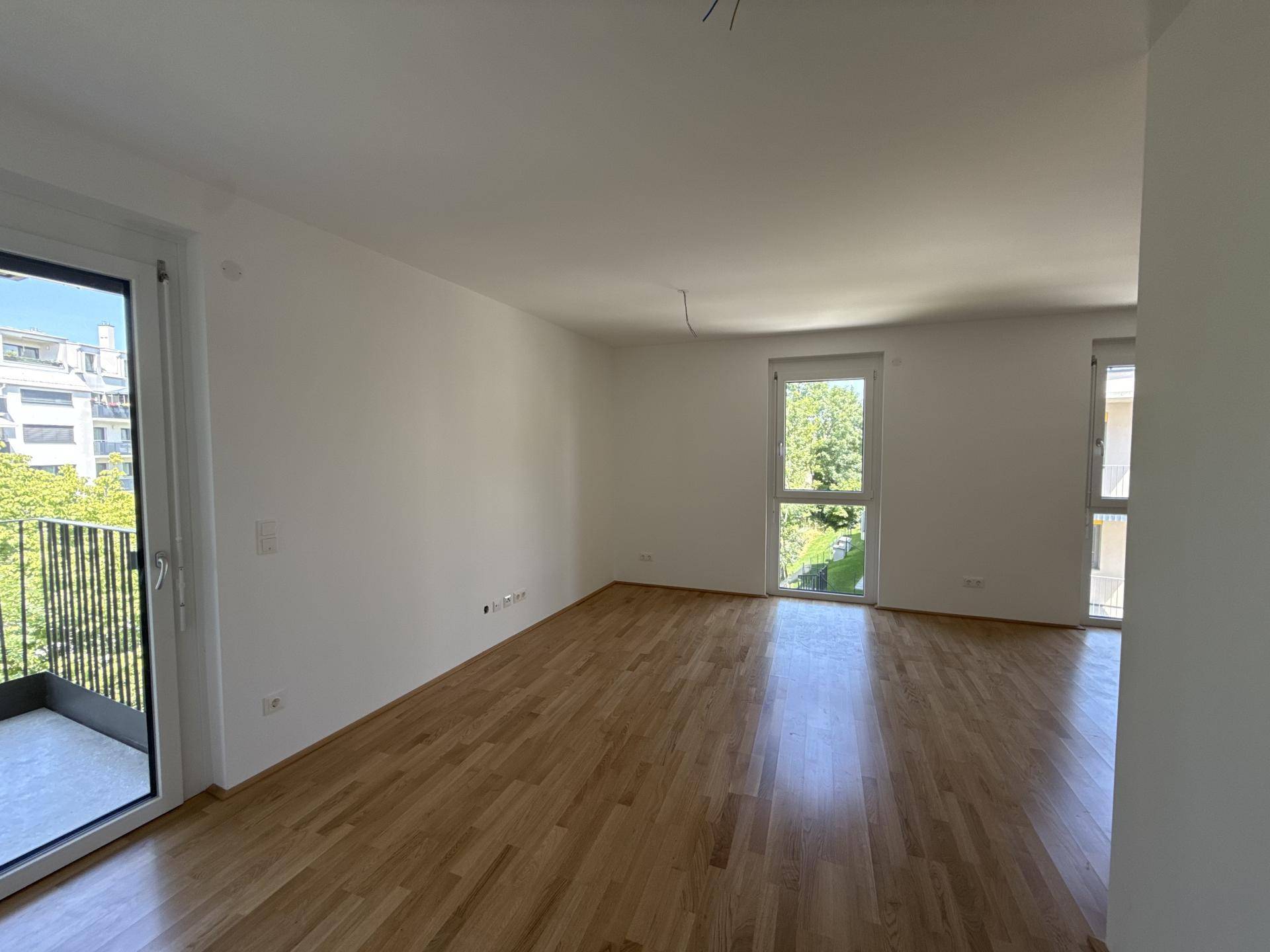 Geräumiger Wohnbereich mit Parkettboden, großen Fenstern und Zugang zum Balkon.