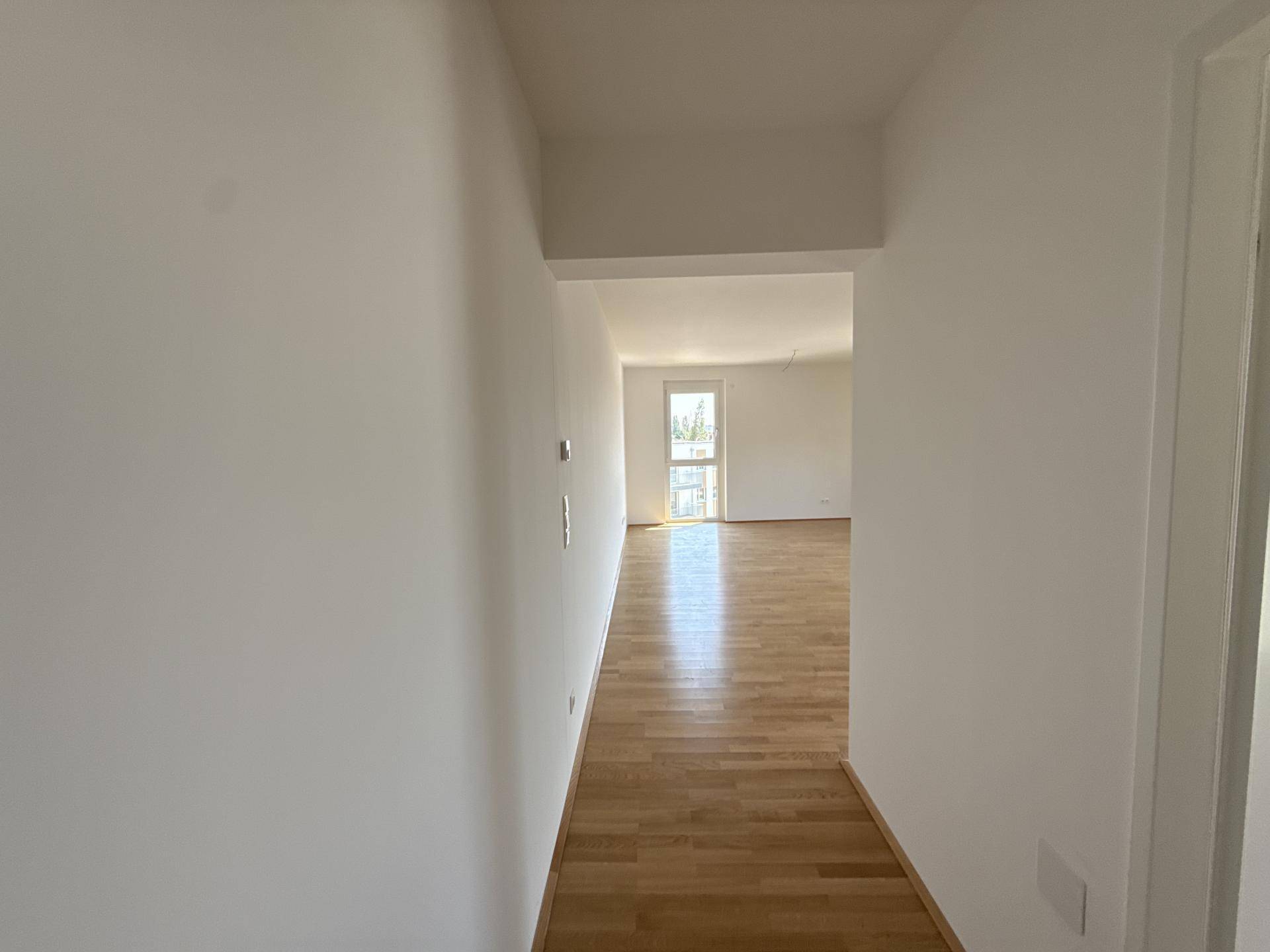 Langer Flur mit Holzparkett, der zu einem hellen Raum mit Fenster führt.