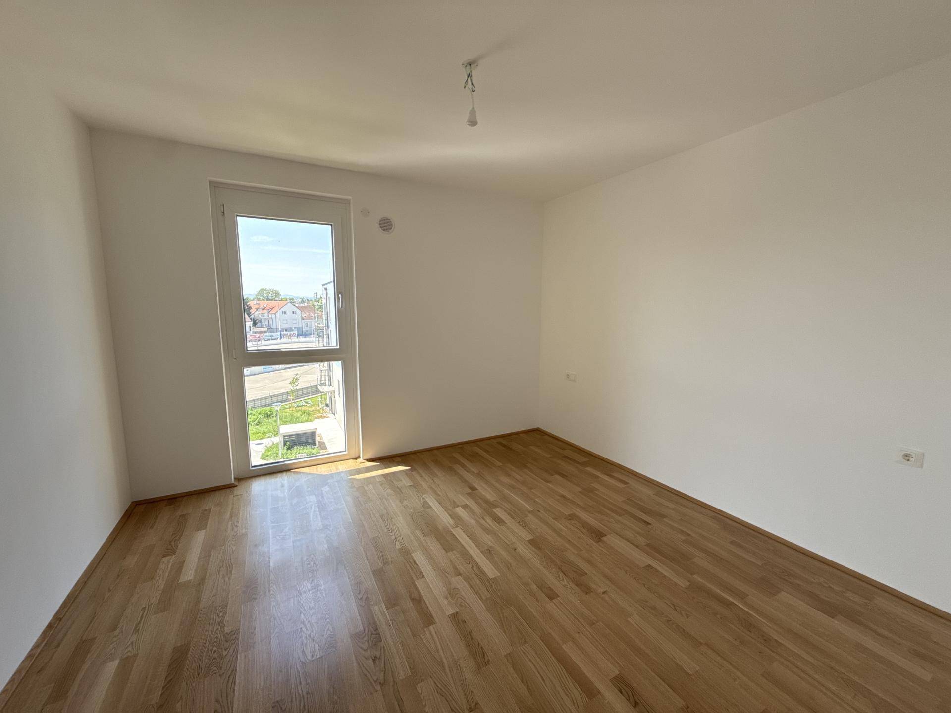 Leeres Schlafzimmer mit hellem Holzparkett und einem großen Fenster für viel Tageslicht.