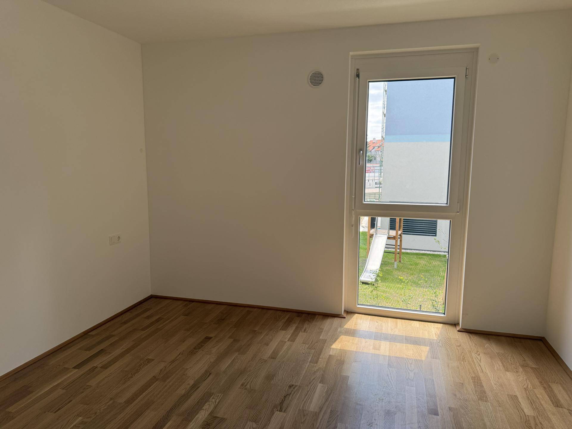 Unmöbliertes Schlafzimmer mit Holzboden und großem Fenster, das auf eine Grünfläche blickt.