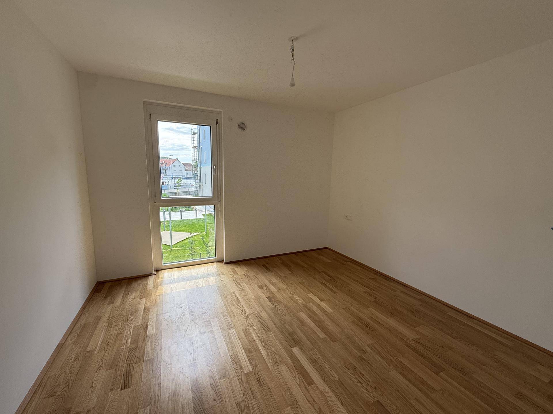 Helles, leeres Schlafzimmer mit neuem Holzparkettboden und großem Fenster mit Blick ins Grüne.