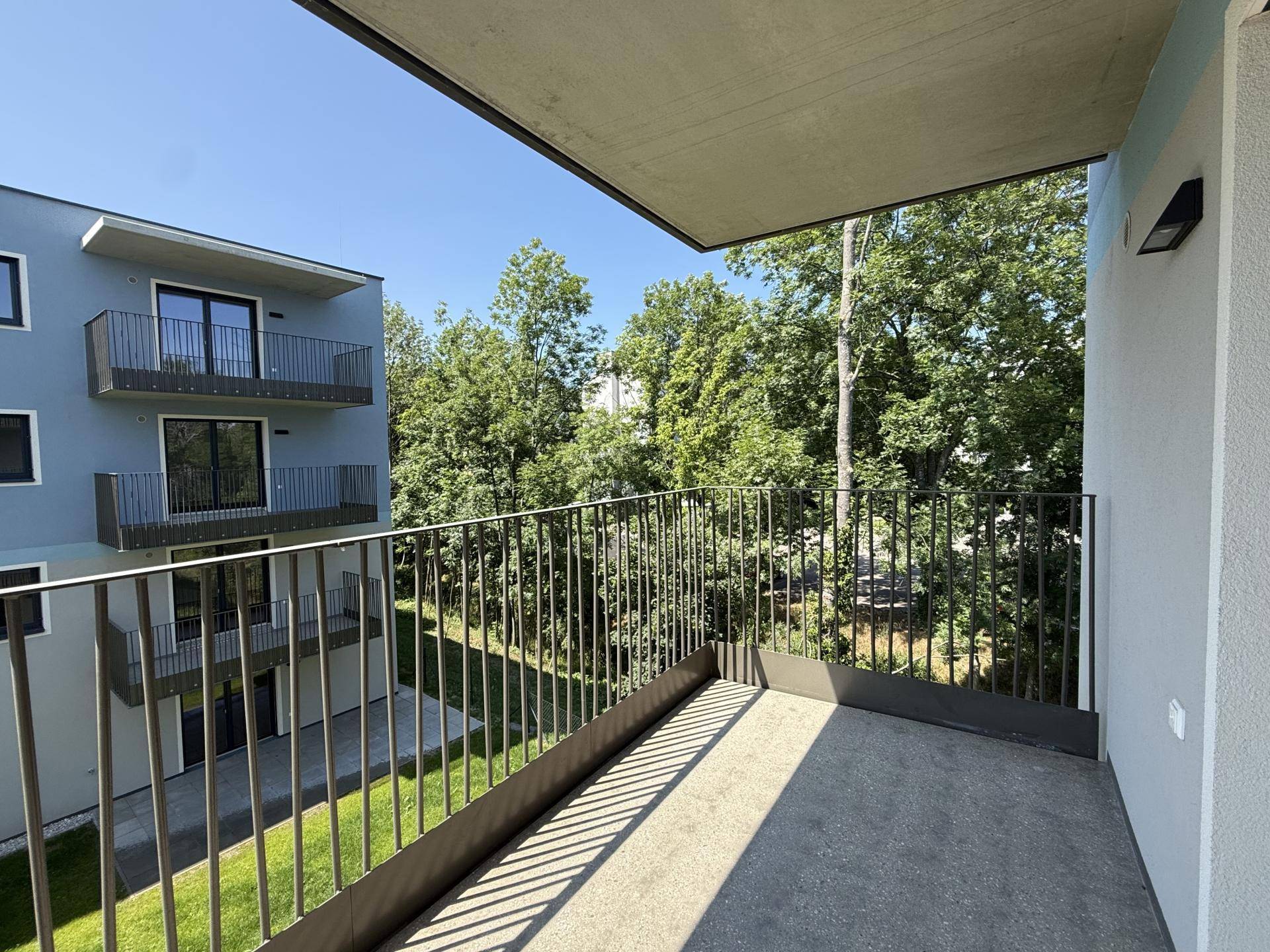 Balkon mit Metallgeländer und Blick auf grüne Bäume und ein angrenzendes Gebäude.