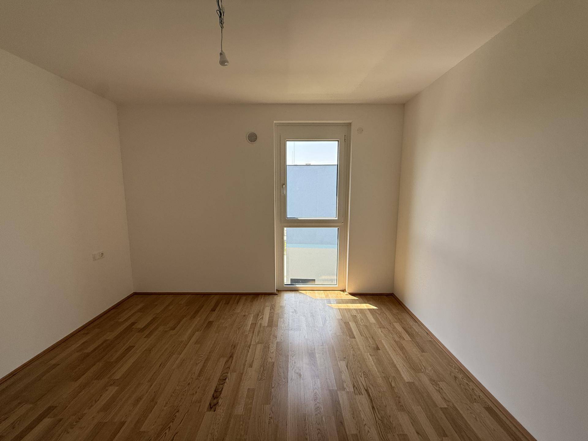Helles, leeres Zimmer mit Holzboden und Fenster, das viel Tageslicht hereinlässt.