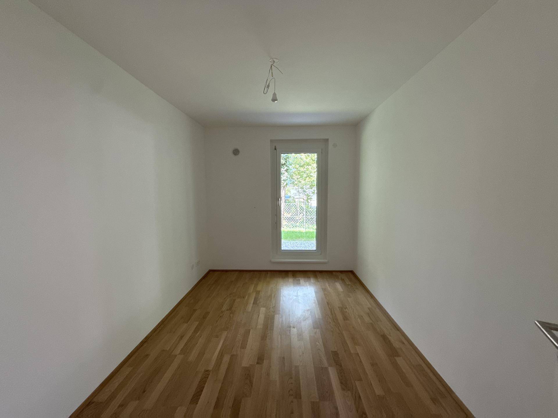 Leeres Zimmer mit Holzboden und einer hohen, schmalen Fenstertür mit Blick ins Grüne.