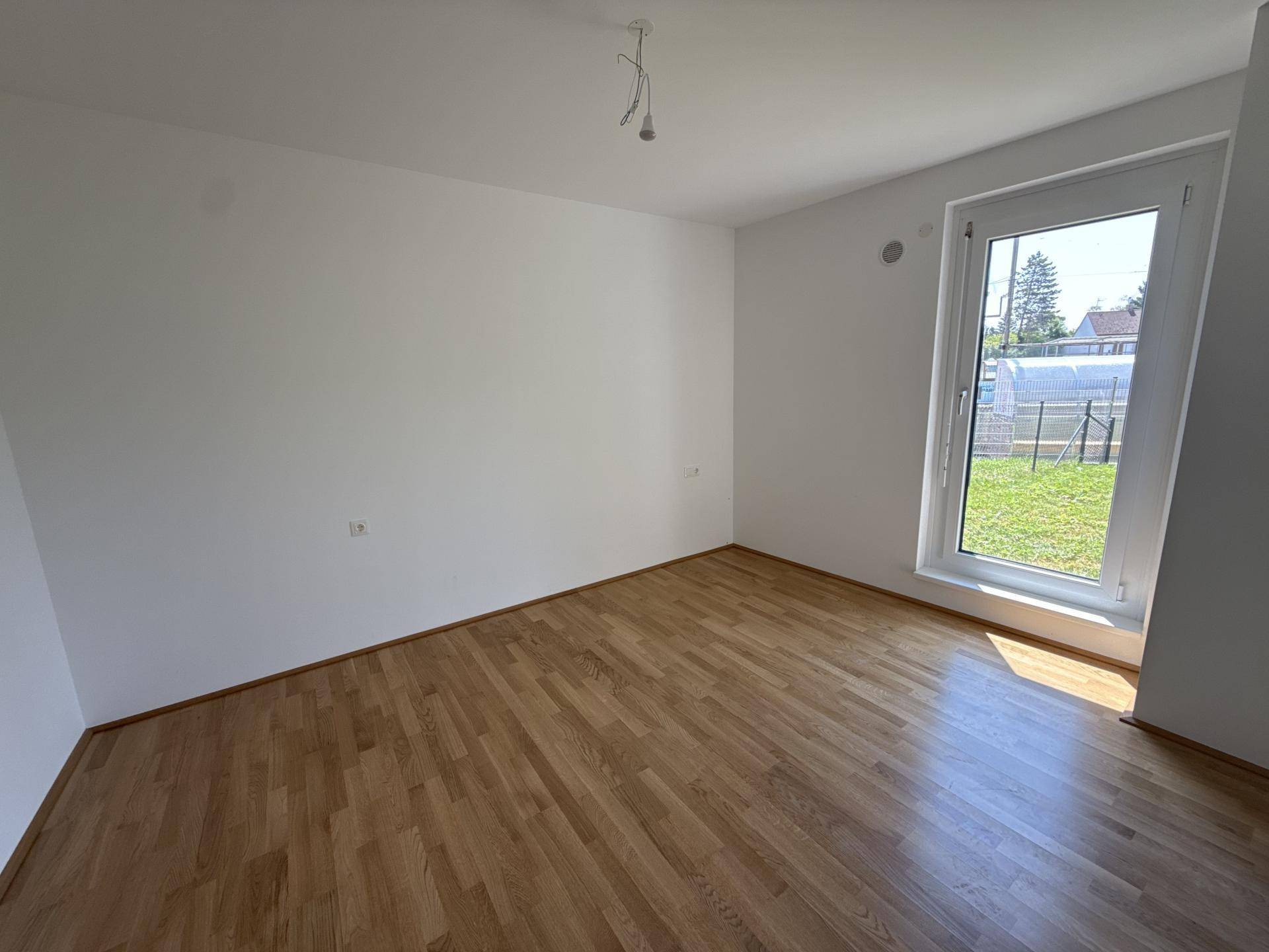 Helles, unmöbliertes Schlafzimmer mit Holzboden und großem Fenster mit Blick ins Grüne.