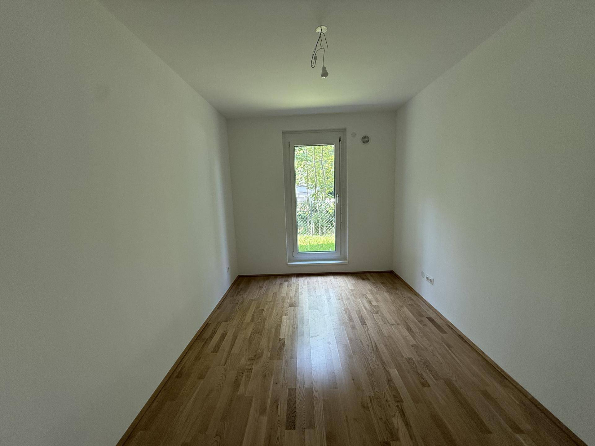 Weiteres helles Schlafzimmer mit Holzparkettboden und einer Fenstertür, die den Blick ins Grüne freigibt.