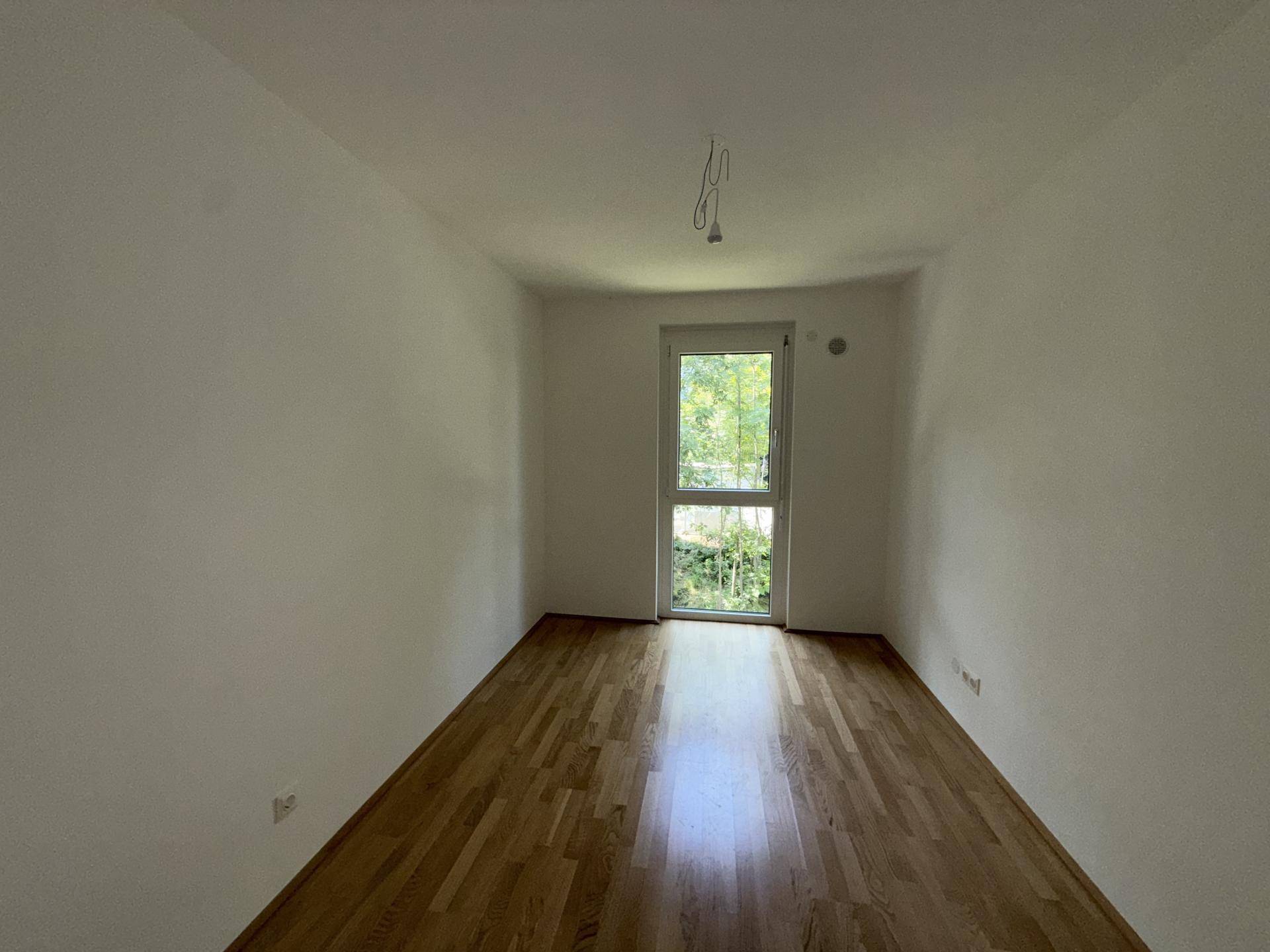 Helles Zimmer mit Holzboden und großem Fenster, das einen Blick ins Grüne bietet.