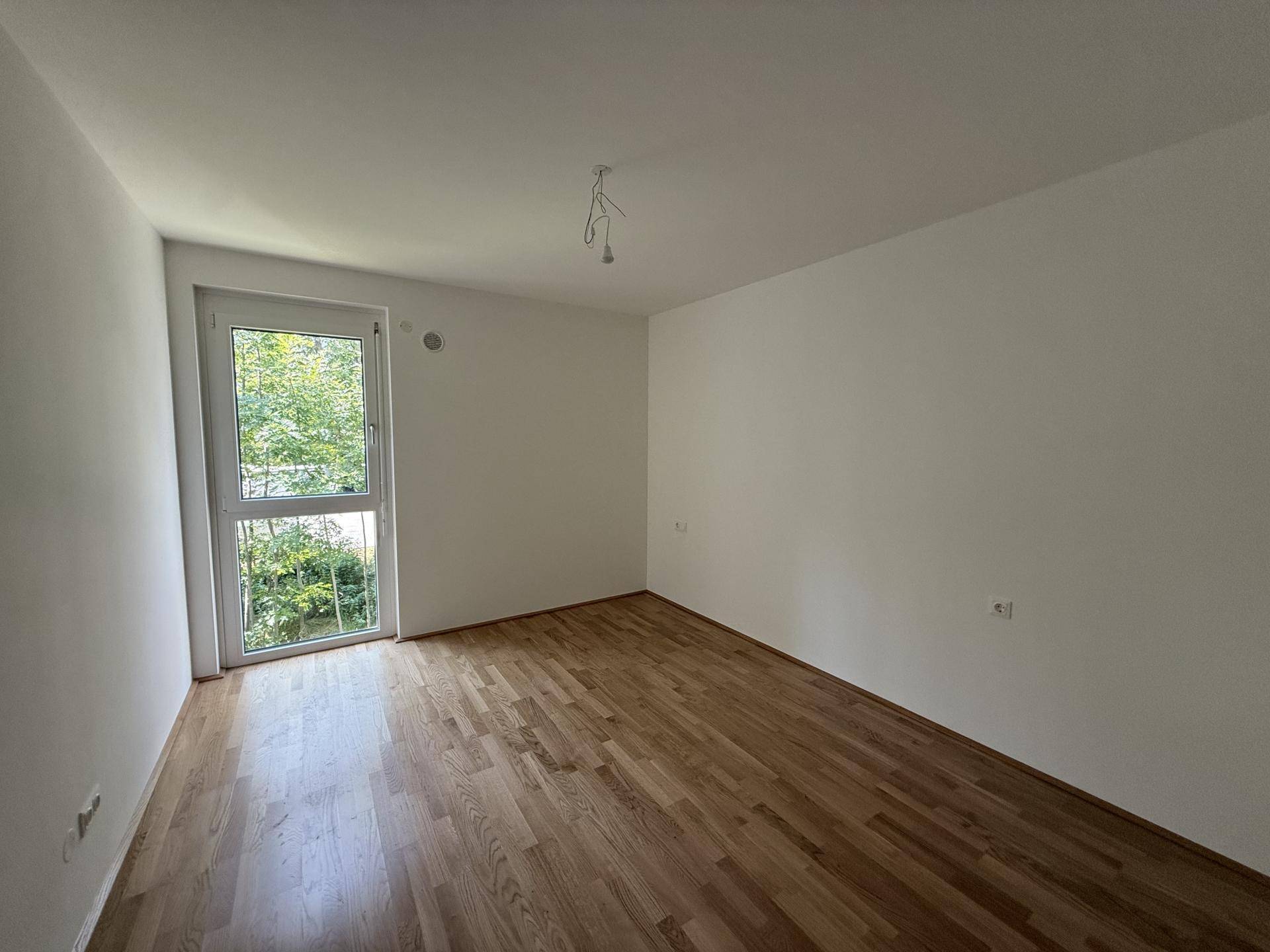 Leeres Zimmer mit Holzboden und Fenster, das viel Tageslicht hereinlässt und eine ruhige Atmosphäre schafft.