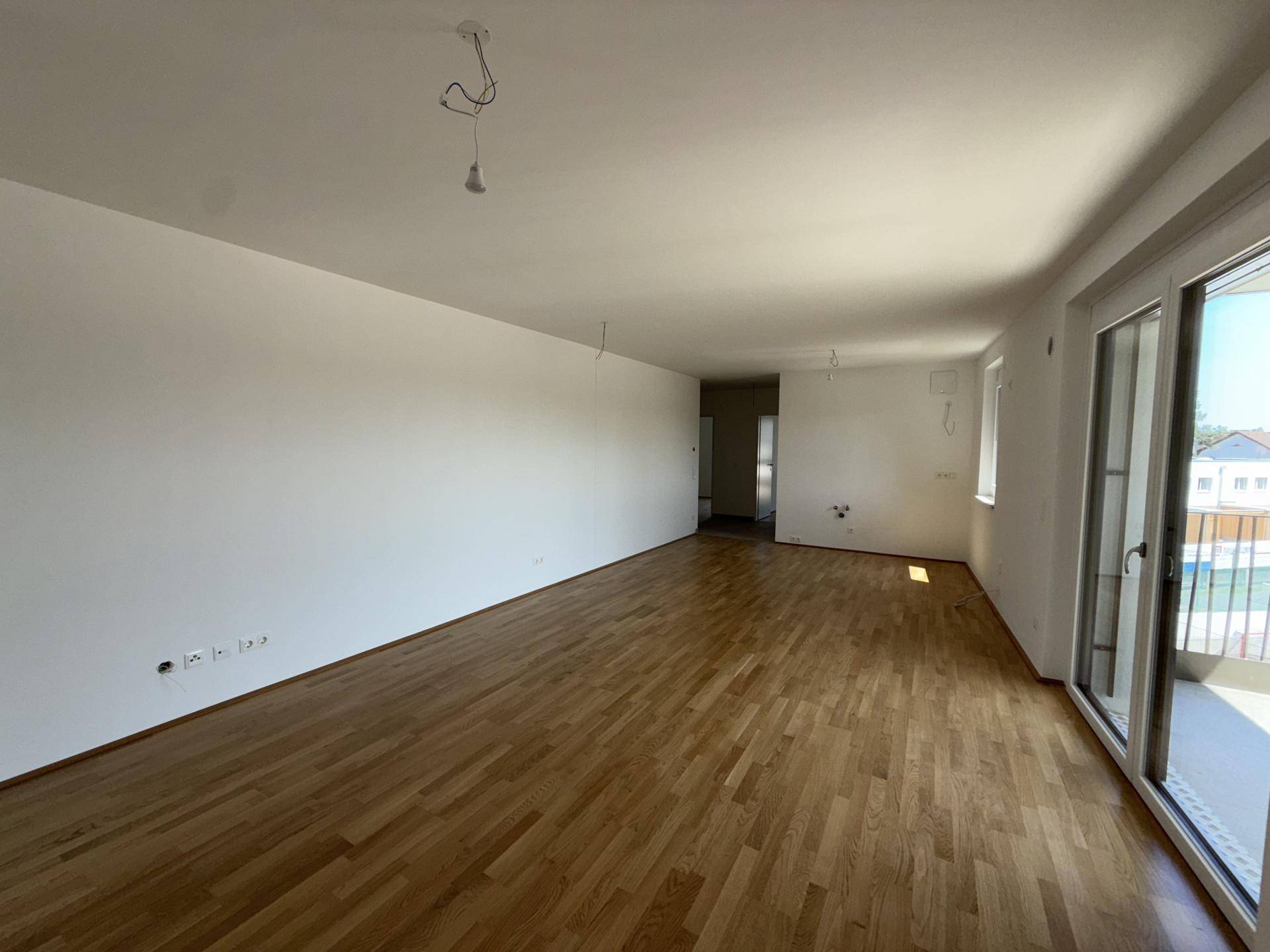 Großzügiges, leeres Wohnzimmer mit hellem Parkettboden und Zugang zu einem Balkon.