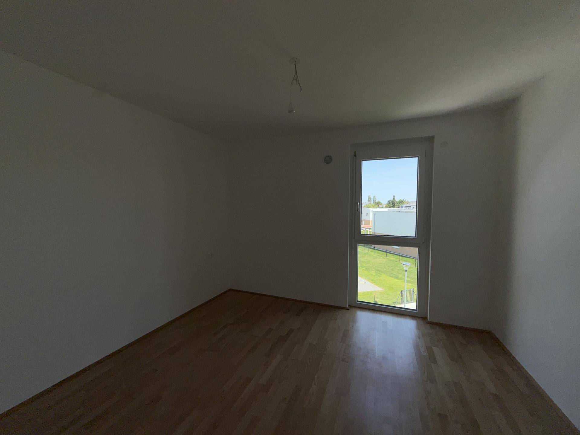 Helles Schlafzimmer mit Parkettboden und einem hohen Fenster, das viel Tageslicht hereinlässt.