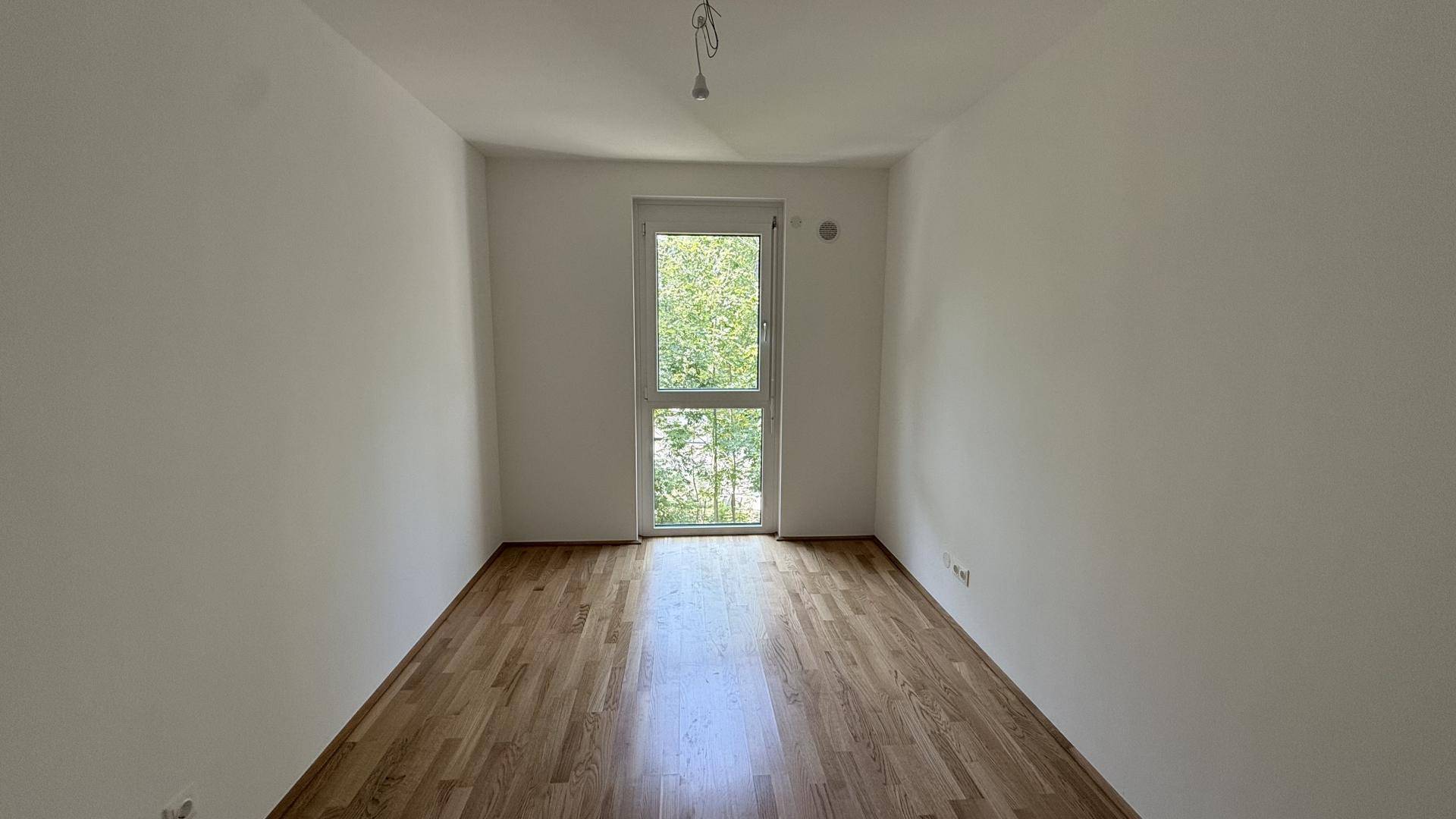 Helles Schlafzimmer mit Holzboden und großem Fenster, das viel Tageslicht hereinlässt und einen grünen Ausblick bietet.