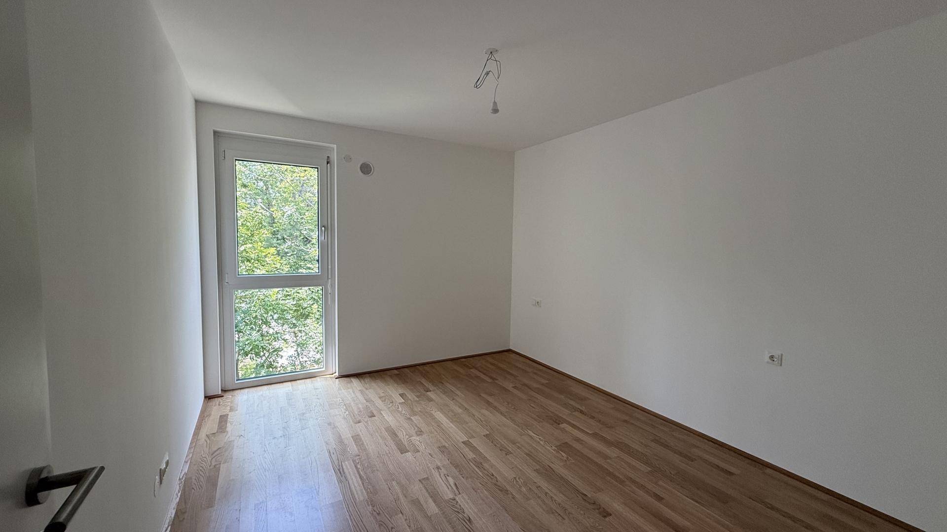 Freundliches Zimmer mit Holzboden und großem Fenster, das einen angenehmen Blick ins Grüne ermöglicht.