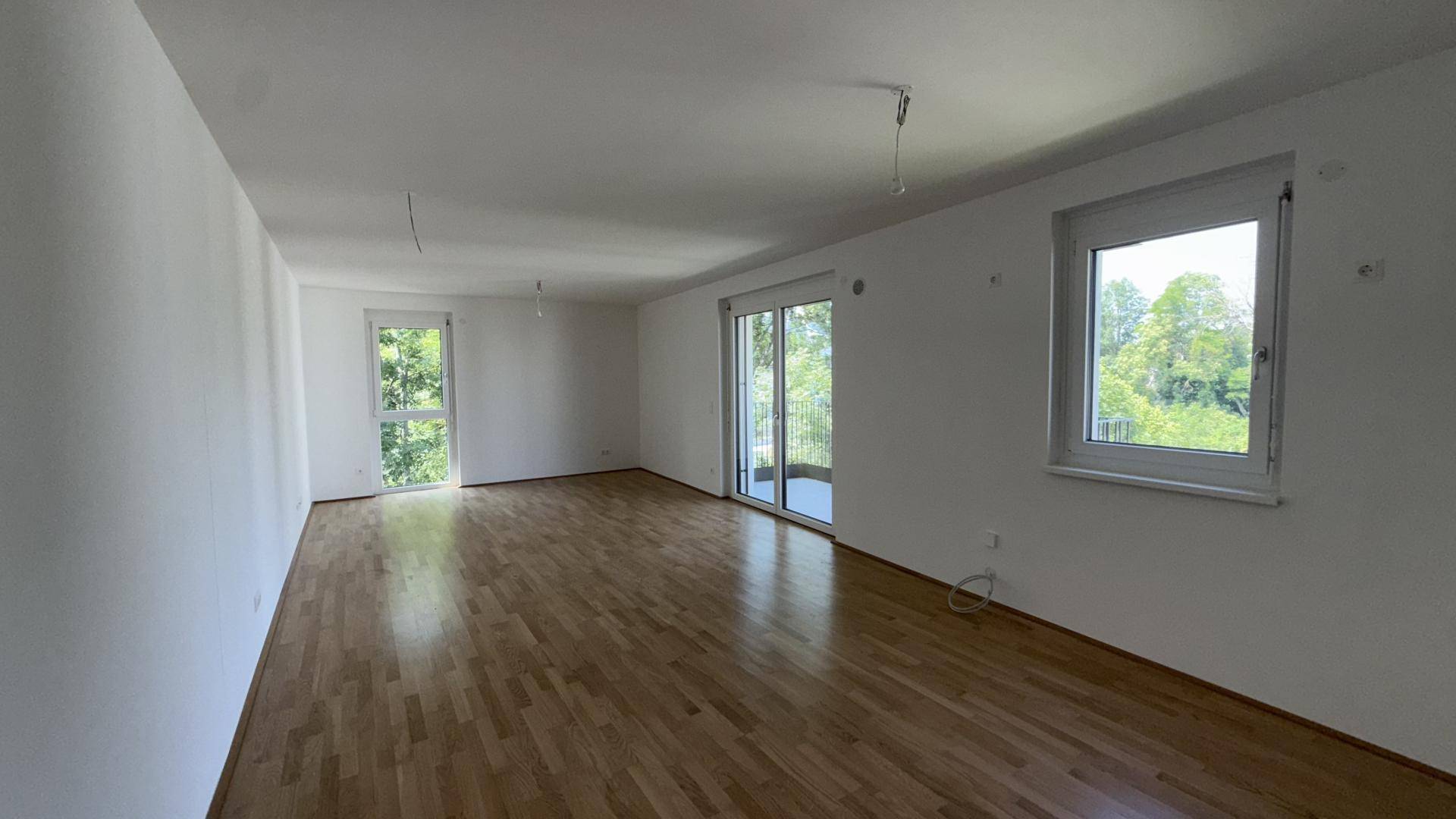 Geräumiges Wohnzimmer mit Parkettboden, großen Fenstern und Balkonzugang mit Grünblick.