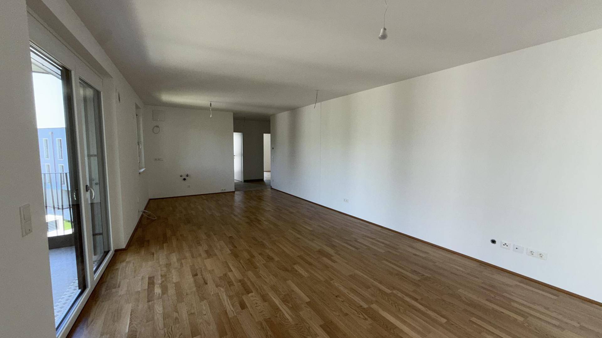 Helles Wohnzimmer mit Holzparkett und Zugang zum Balkon sowie zum offenen Flurbereich.