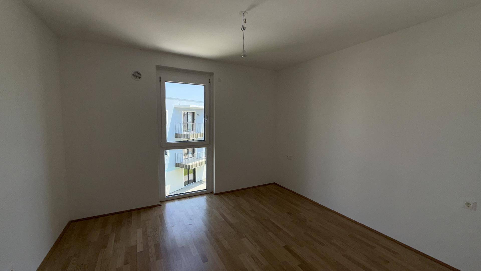 Schlafzimmer mit Parkettboden und großem Fenster, das viel Tageslicht hereinlässt und Blick auf ein Nachbargebäude bietet.