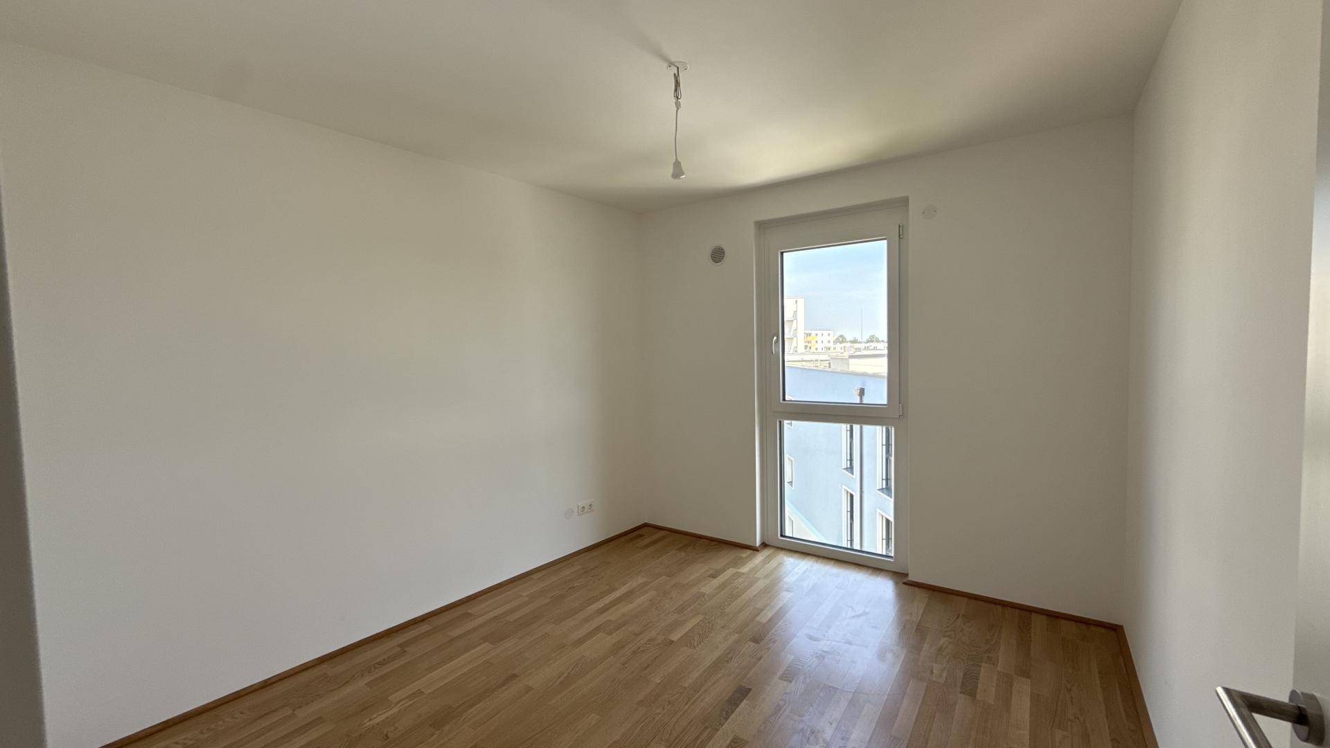 Helles Schlafzimmer mit Holzboden und großem Fenster, das viel Tageslicht hereinlässt.
