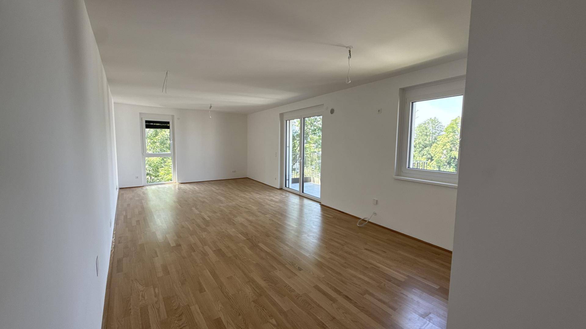 Großes, leeres Wohnzimmer mit hellem Holzparkett und Zugang zu einem sonnigen Balkon.