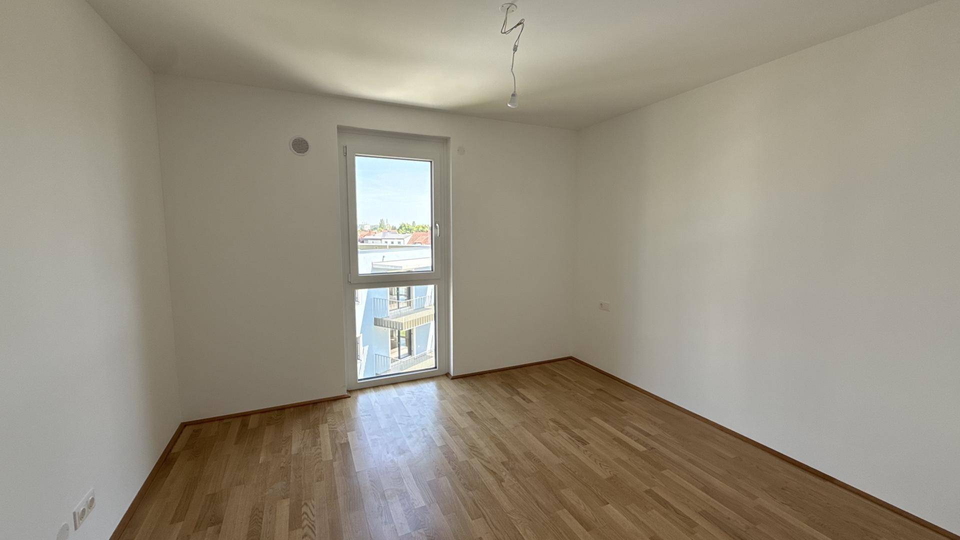 Leeres Schlafzimmer mit hellem Parkettboden und einem großen Fenster für natürliche Belichtung.
