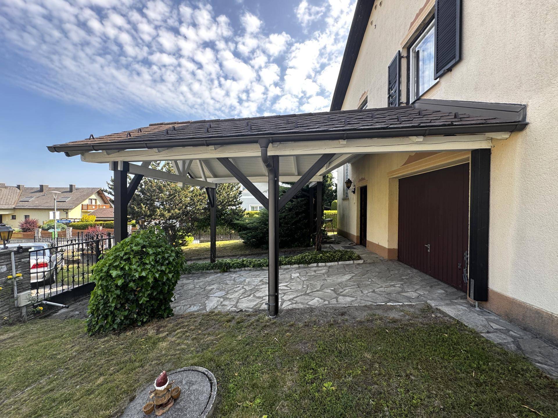 Überdachter Stellplatz (Carport) neben dem Haus mit Zufahrt und Gartenbereich.