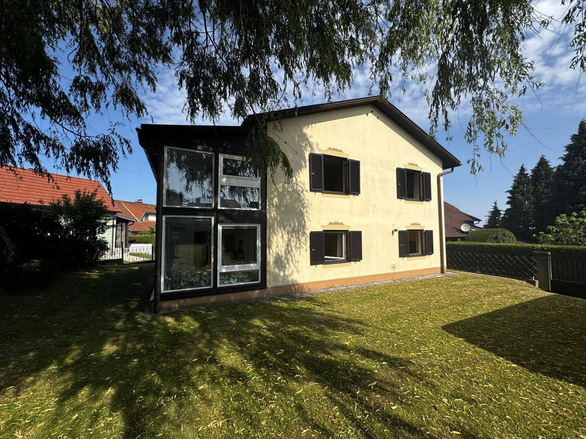 Hausfassade mit großem Wintergarten und gepflegtem Rasen unter Bäumen.