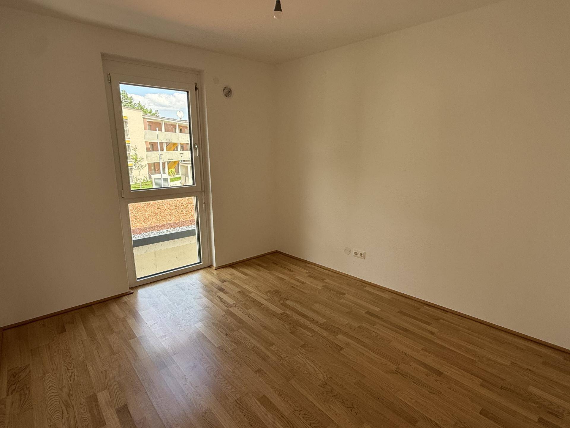 Schlafzimmer mit hellem Parkettboden, weißen Wänden und einem großen Fenster mit Blick ins Grüne.