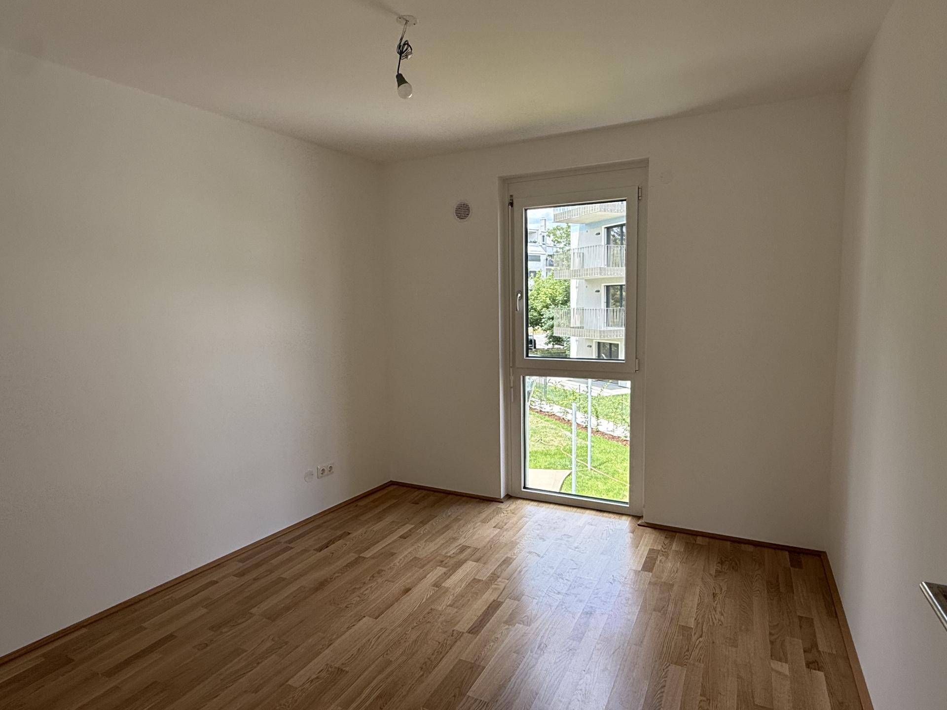 Weiteres helles Schlafzimmer mit Holzboden und großem Fenster, das viel Tageslicht hereinlässt.