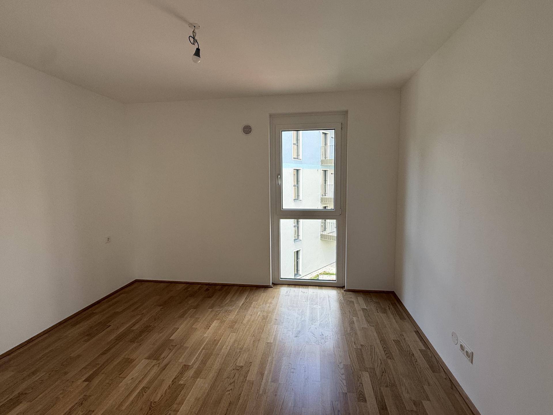 Weiteres helles Zimmer mit Parkettboden und einem hohen Fenster mit Blick auf die Umgebung.
