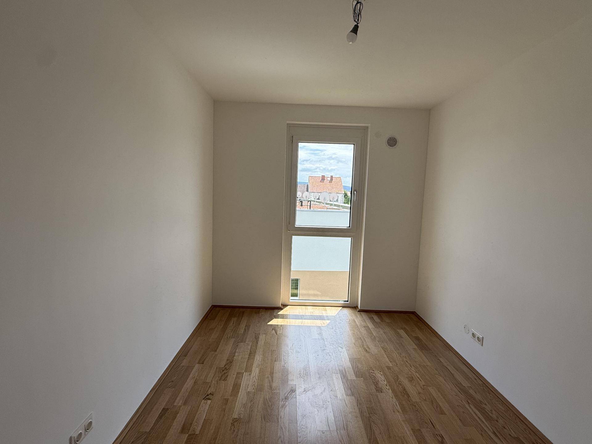 Helles Zimmer mit Holzboden und einem großen Fenster, das viel Tageslicht hereinlässt.