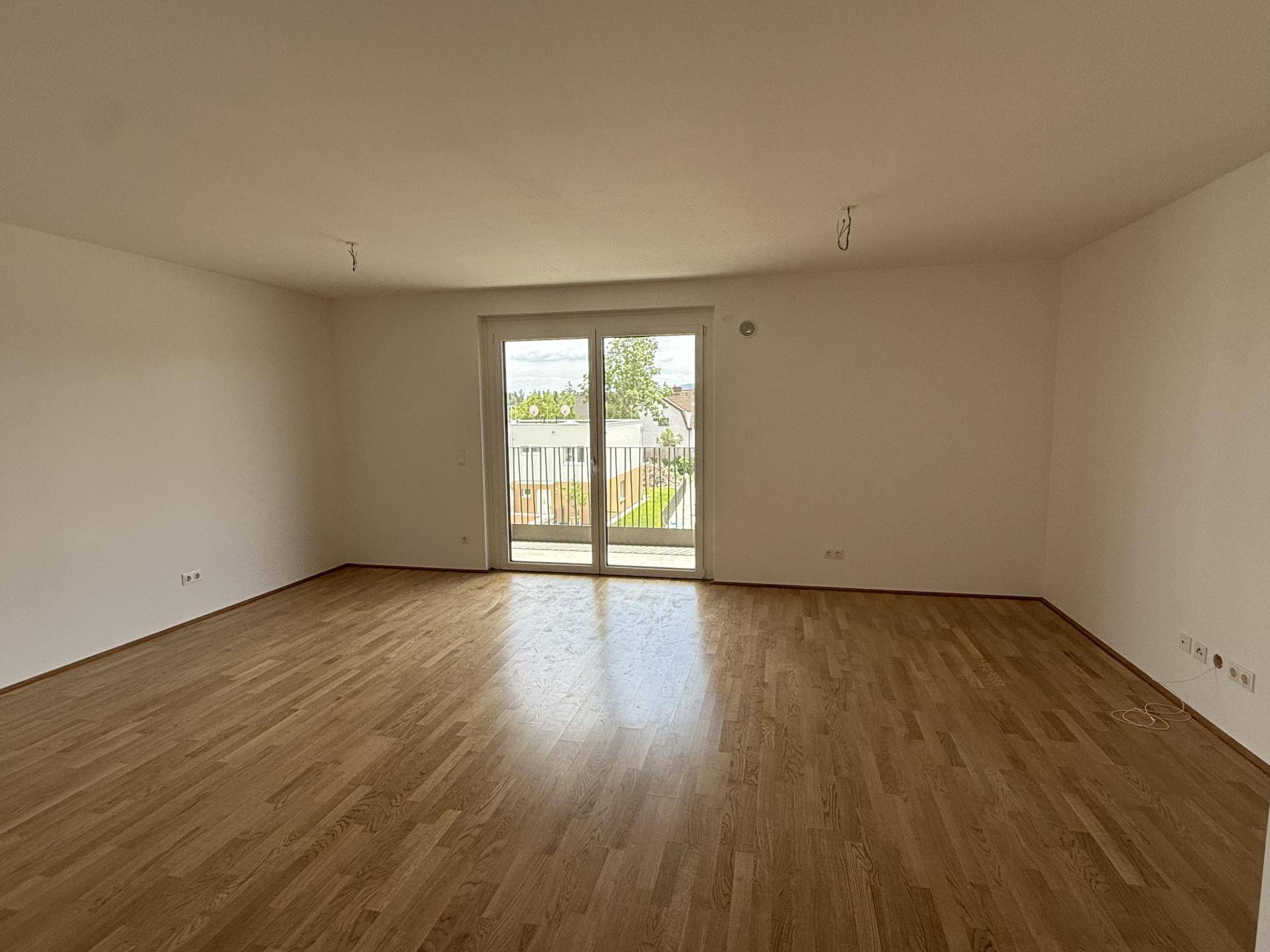Geräumiges, leeres Wohnzimmer mit hellem Parkettboden und großem Fenster mit Balkonzugang.