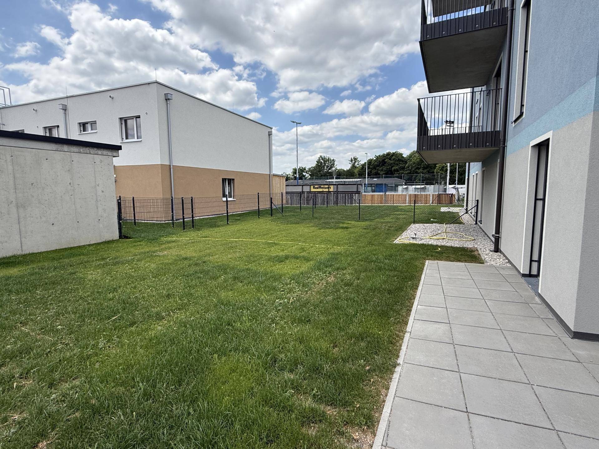 Weitläufige Rasenfläche neben dem Neubau mit gepflastertem Weg und Zaun.