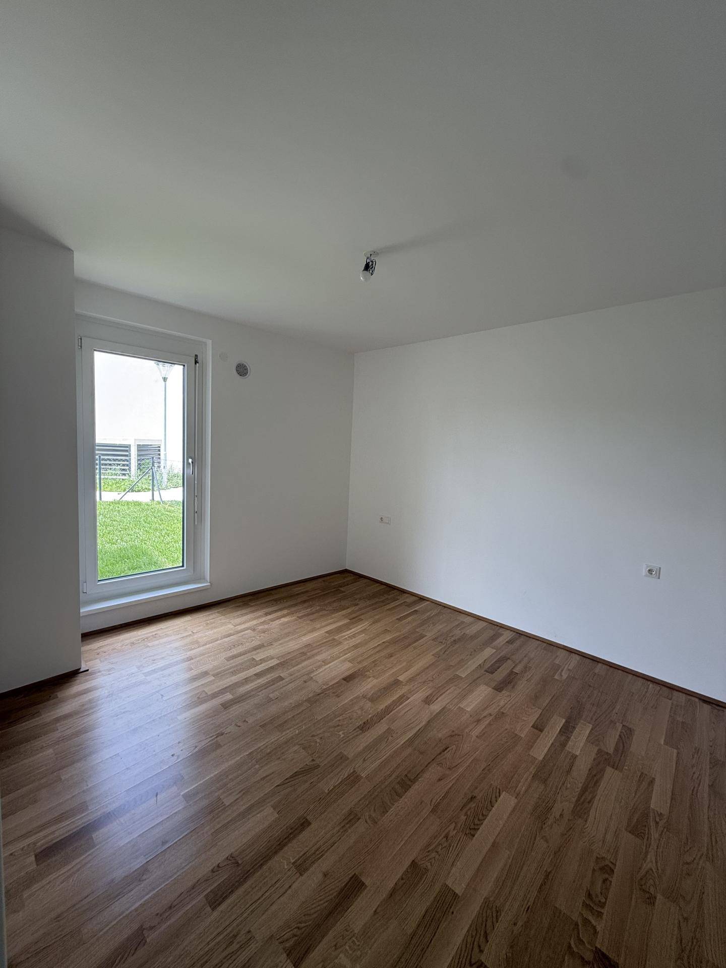 Lichtdurchflutetes Zimmer mit Holzparkett und einer Fenstertür, die den Blick ins Grüne freigibt.