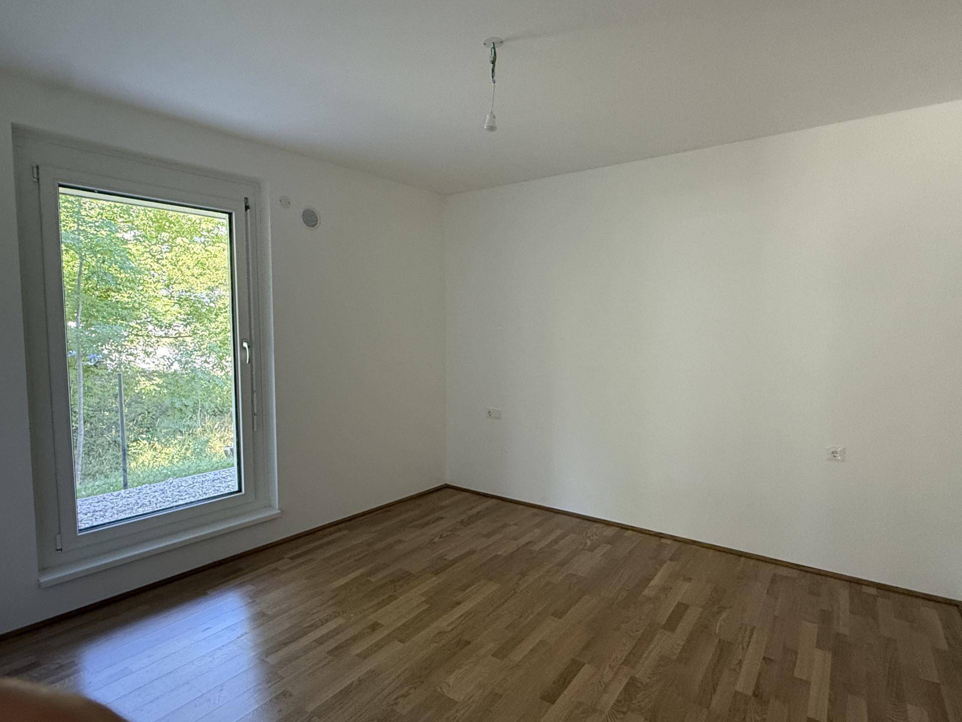 Helles Zimmer mit Parkettboden und großem Fenster, das einen angenehmen Blick auf Bäume bietet.