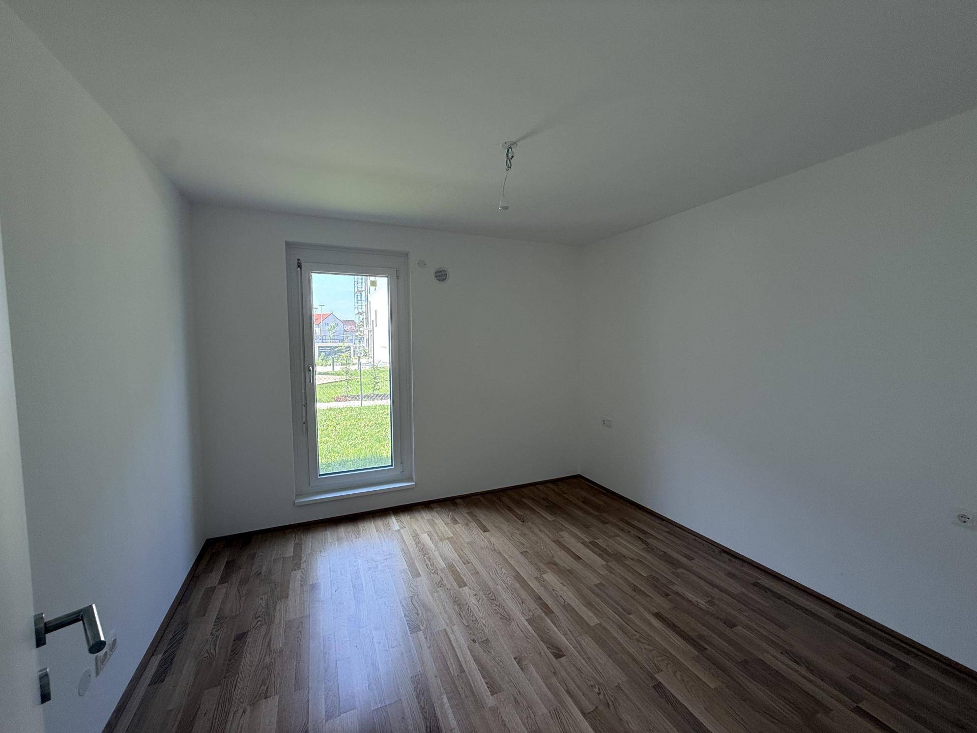 Leeres Schlafzimmer mit Holzboden und großem Fenster mit Blick ins Grüne.