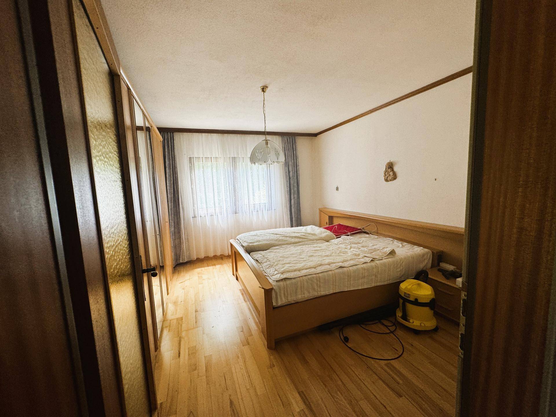 Schlafzimmer mit Doppelbett, Holzschrank und Parkettboden, hell durch großes Fenster.