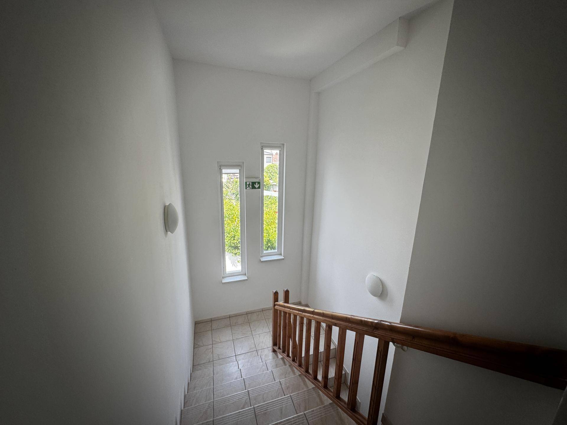 Treppenhaus mit Holzgeländer und Fliesenboden, beleuchtet durch hohe, schmale Fenster.