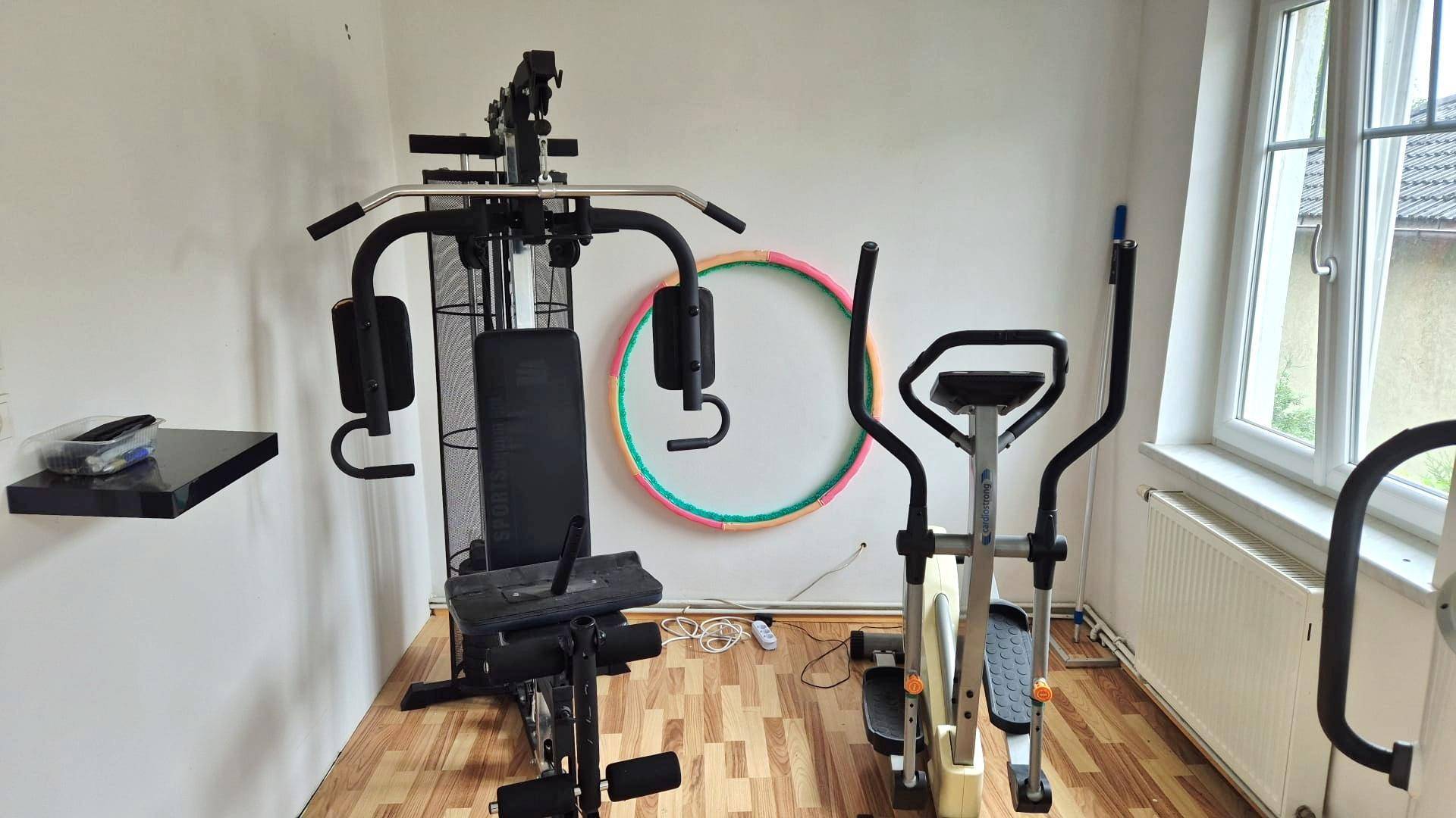 Fitnessraum mit verschiedenen Trainingsgeräten und Holzboden.