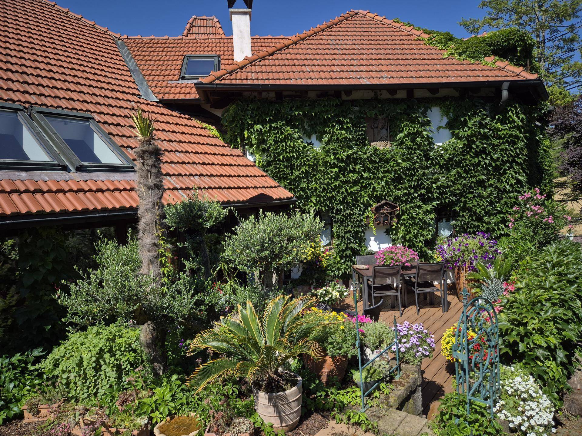 Malerischer Garten mit üppiger Vegetation, Terrasse und traditionellem Haus mit Ziegeldach.