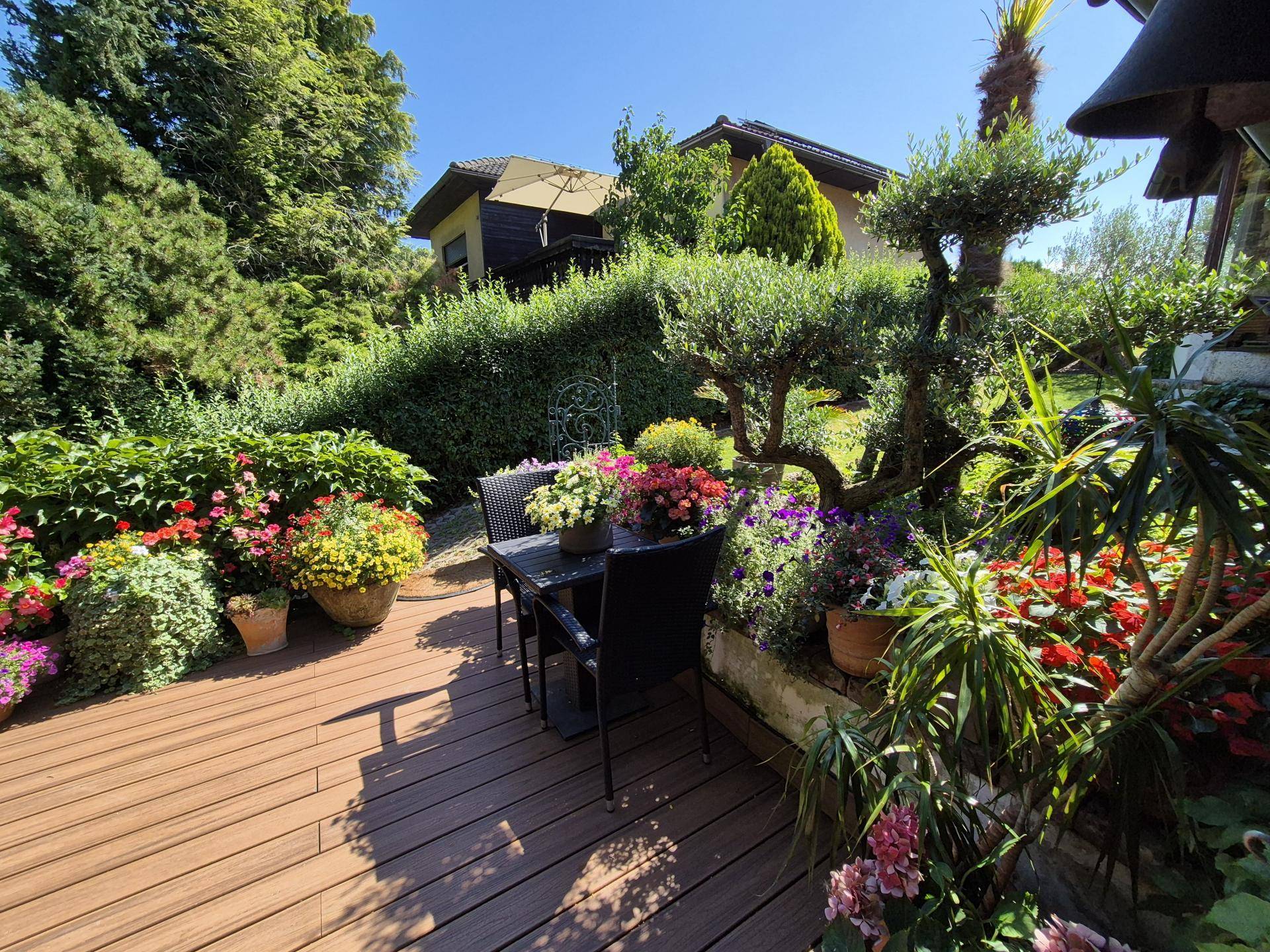 Sonnige Holzterrasse mit Gartenmöbeln, umgeben von blühenden Pflanzen und Bäumen.