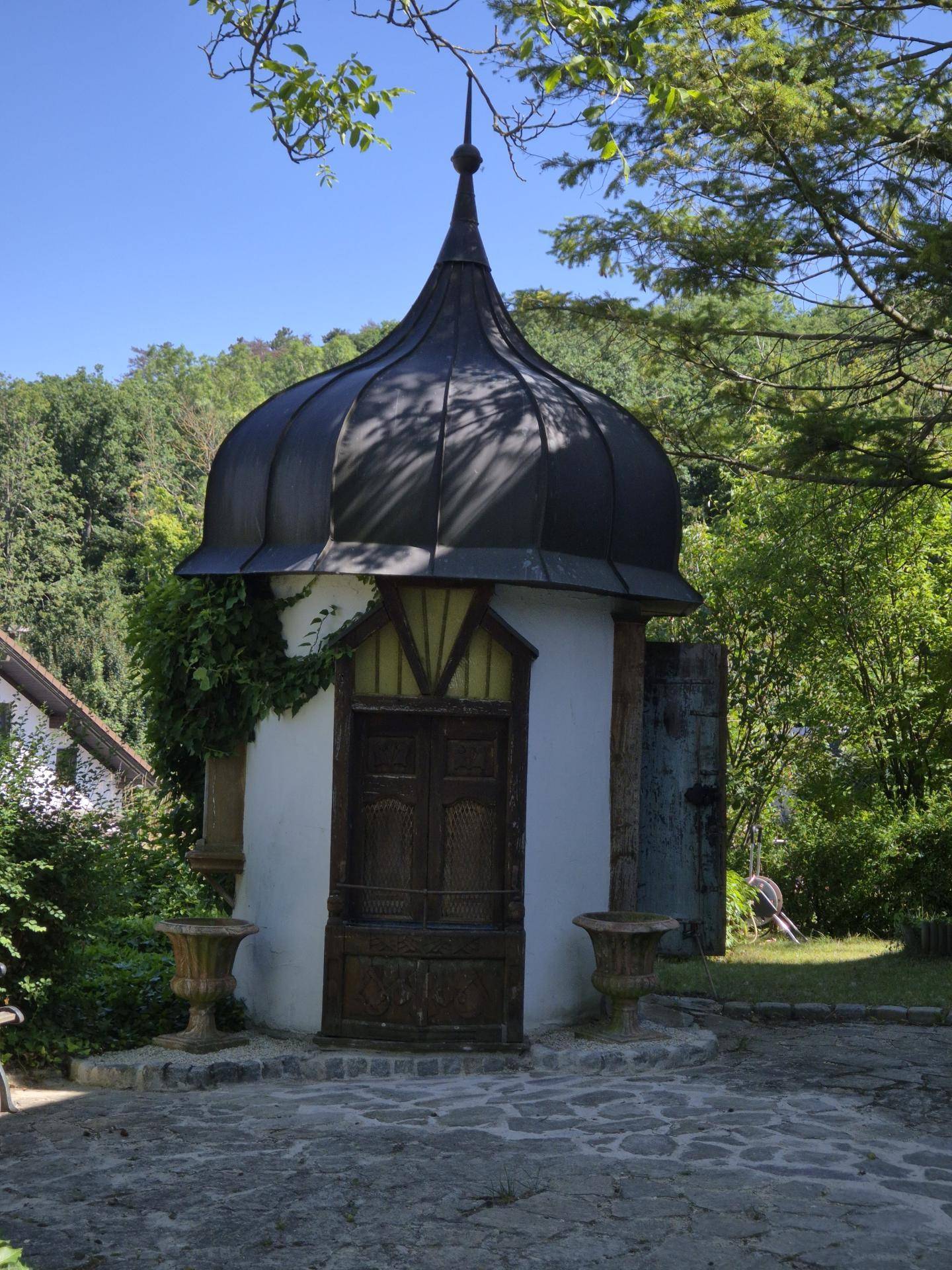 Kleines, dekoratives Gebäude mit Kuppeldach und Holztür in einer grünen Umgebung.