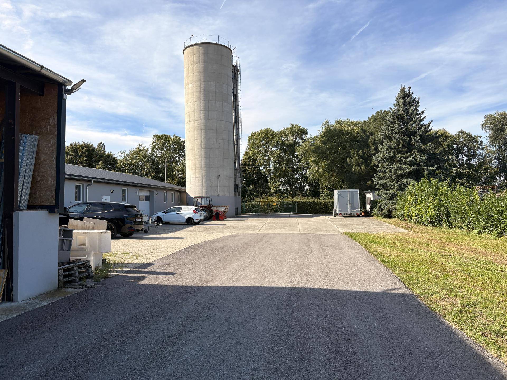 Blick auf das Gelände mit einem großen Silo, einem Gebäude und einem asphaltierten Parkplatz unter blauem Himmel.