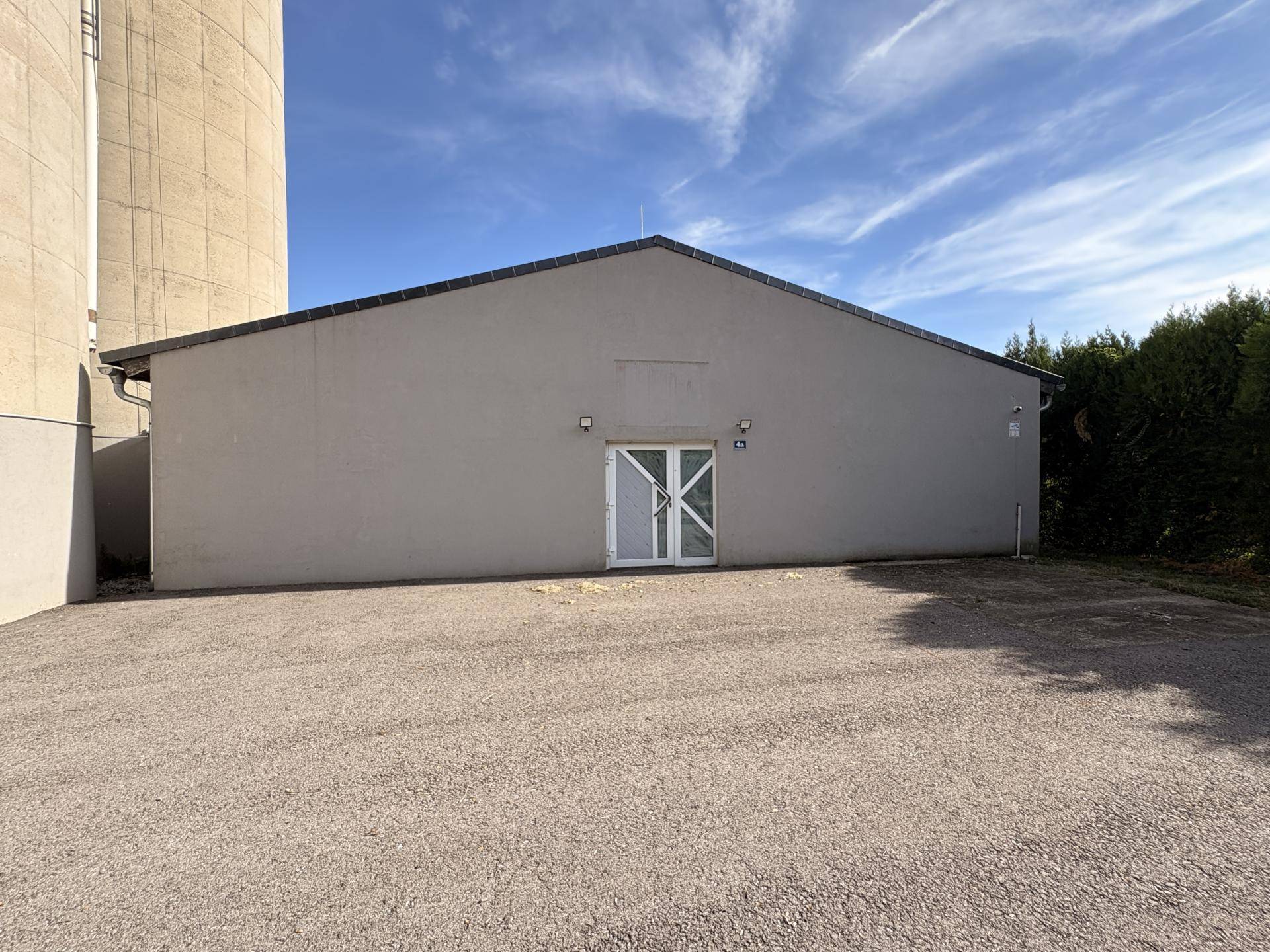 Rückseite eines grauen Gebäudes mit Doppeltür und einem Kiesbereich, neben einem großen Silo.