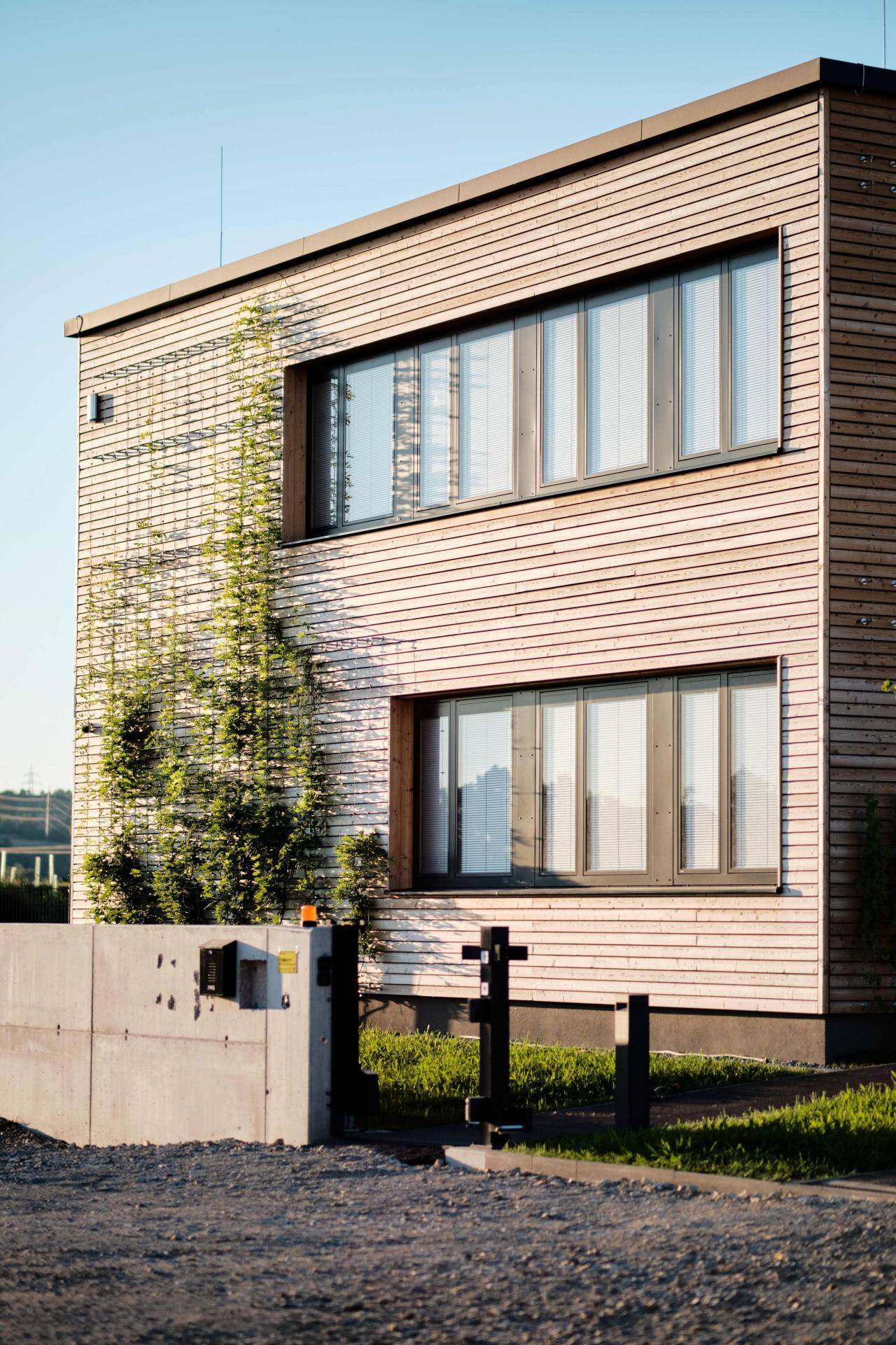 Seitenansicht des Gebäudes mit Holzfassade, großen Fenstern und einem modernen Zugangstor.