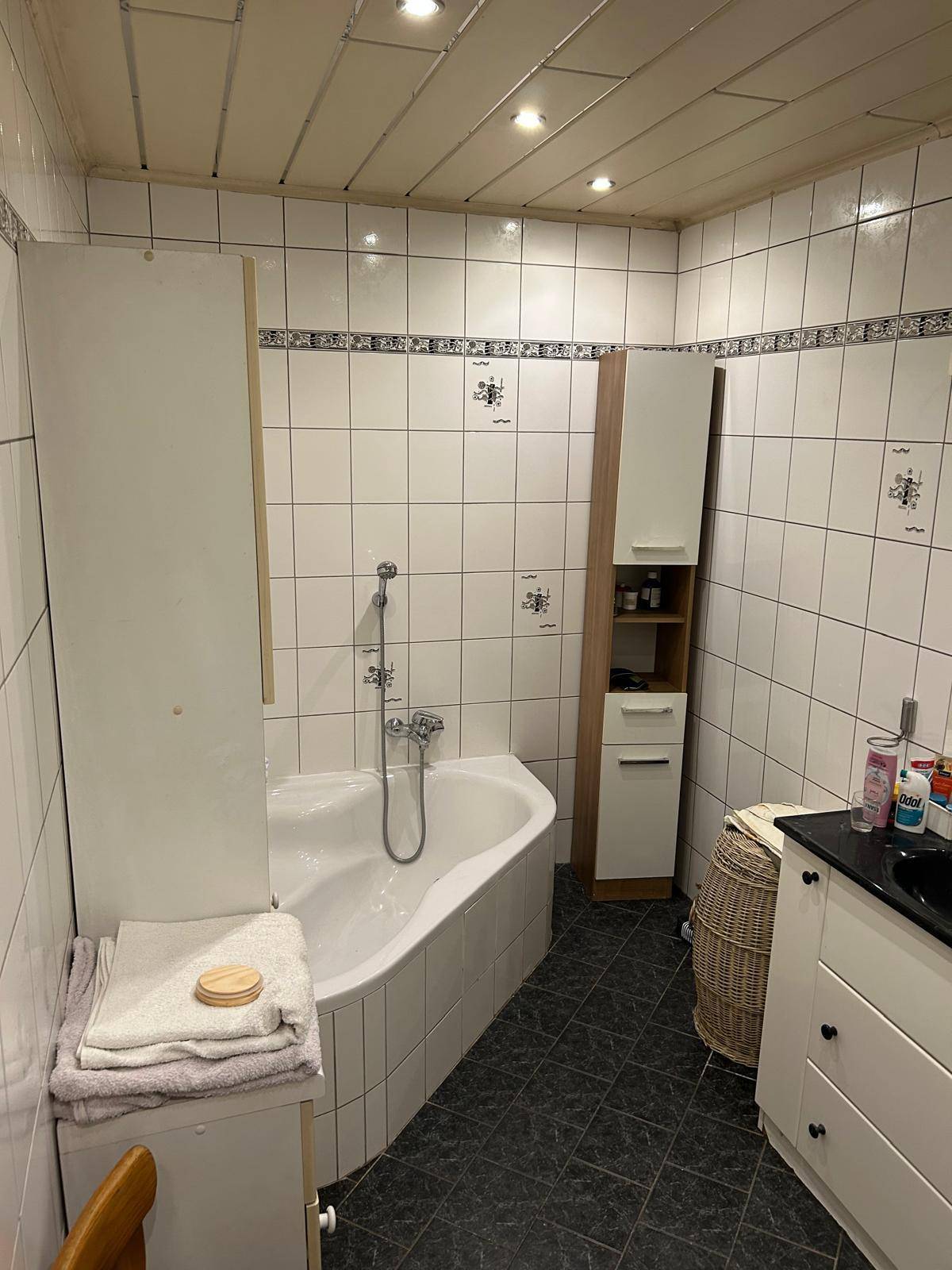 Badezimmer mit weiß gefliesten Wänden, Eckbadewanne und dunklem Fliesenboden.