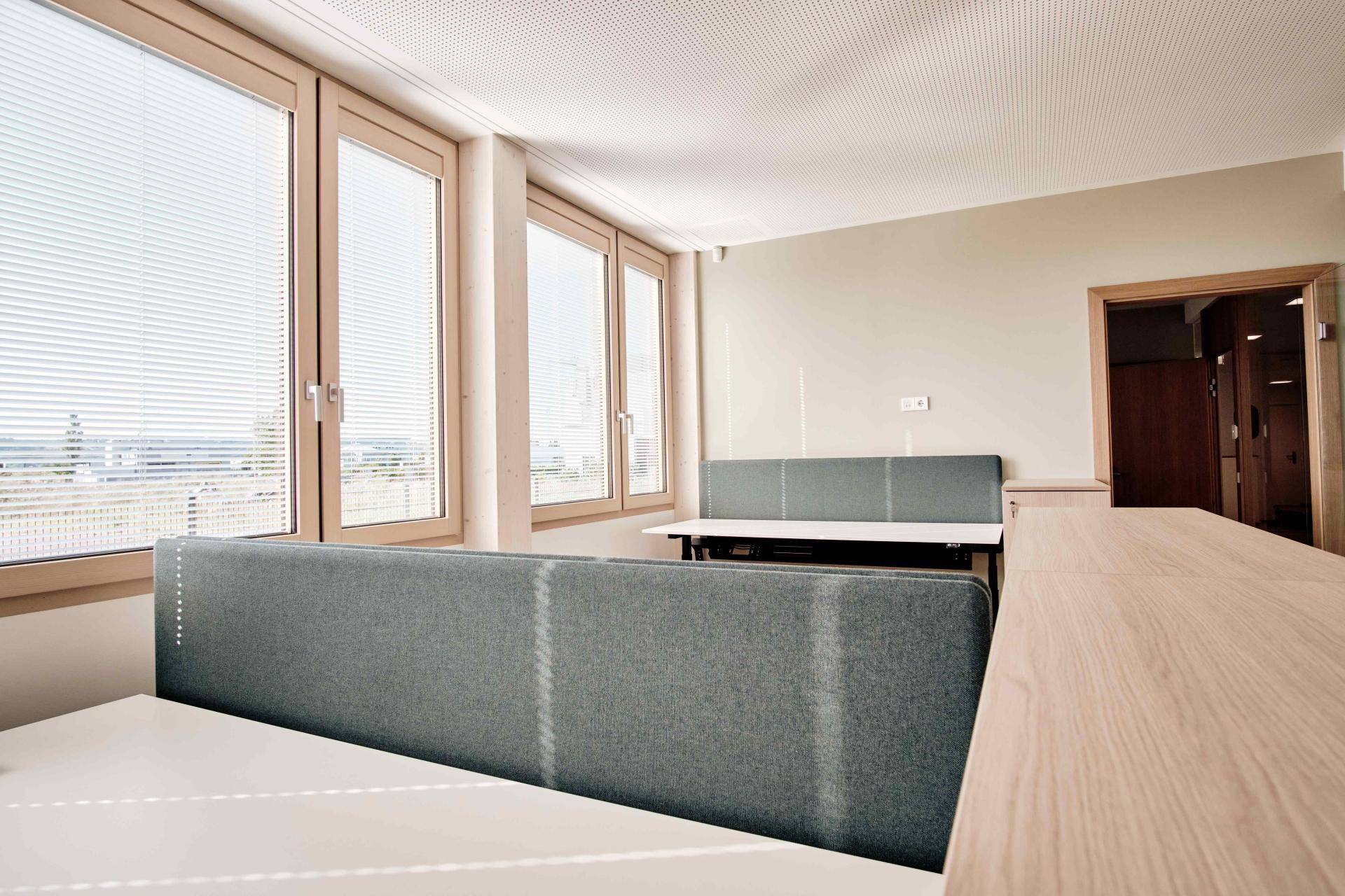 Heller Büroraum mit mehreren Arbeitsplätzen, großen Fenstern und Holzrahmen.
