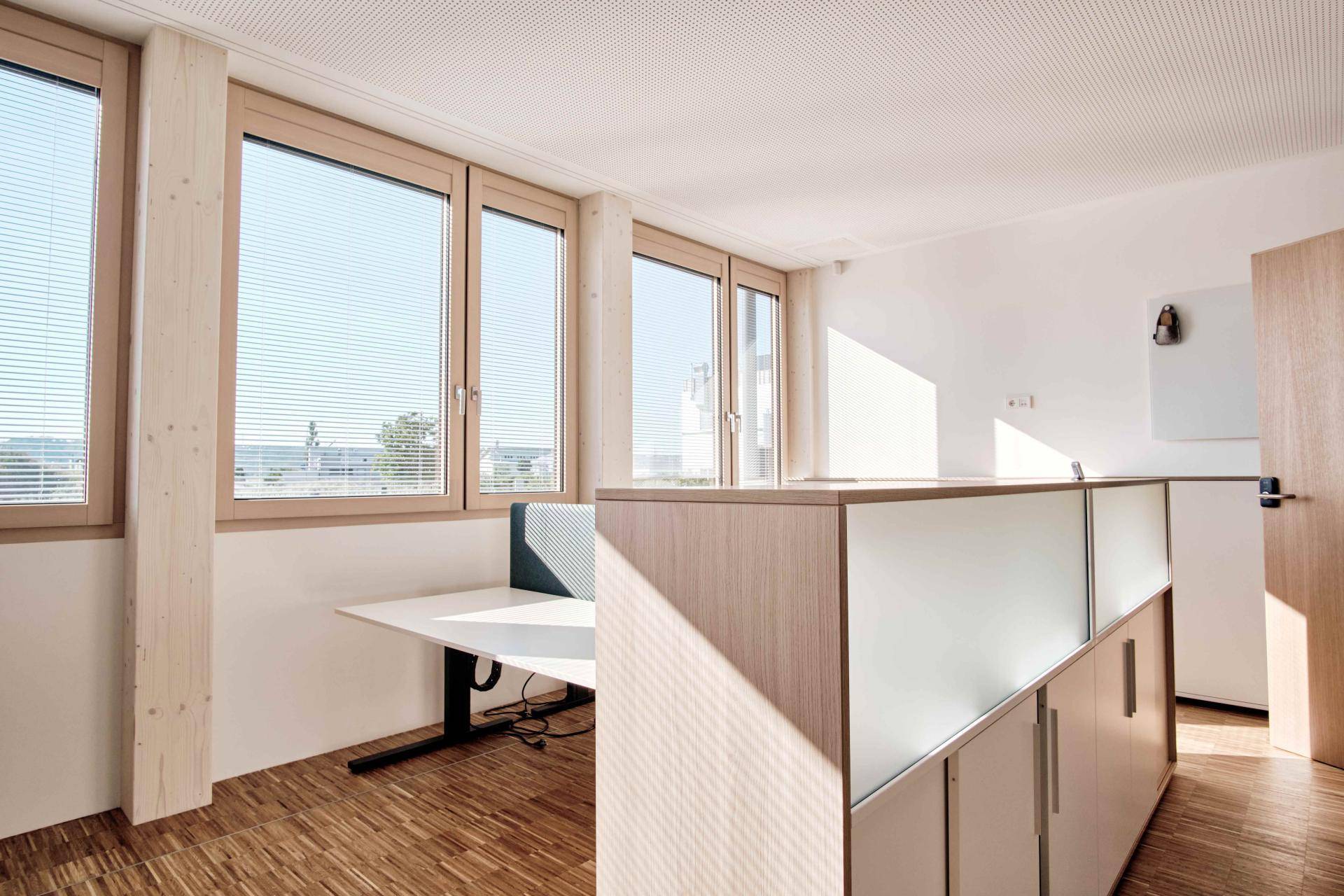 Heller Büroraum mit modernen Arbeitsplätzen, großen Fenstern und Holzfußboden.