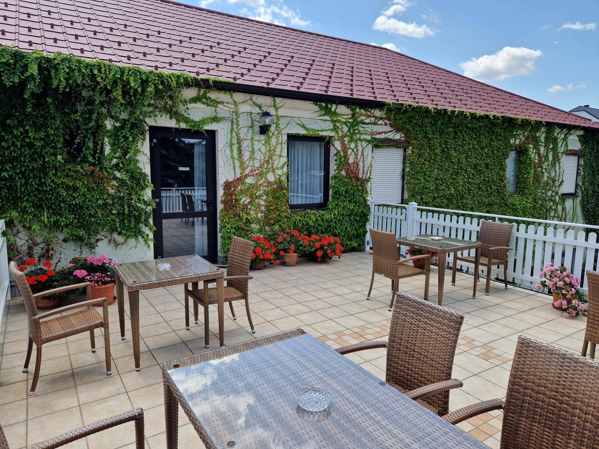 Gemütliche Terrasse mit Korbmöbeln und Pflanzen, umgeben von grüner Fassade und weißem Zaun.