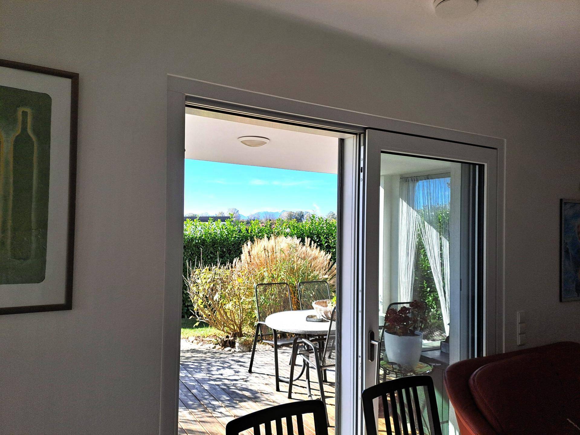 Blick vom Innenraum auf die sonnige Holzterrasse mit Gartenmöbeln und Bergblick in der Ferne.