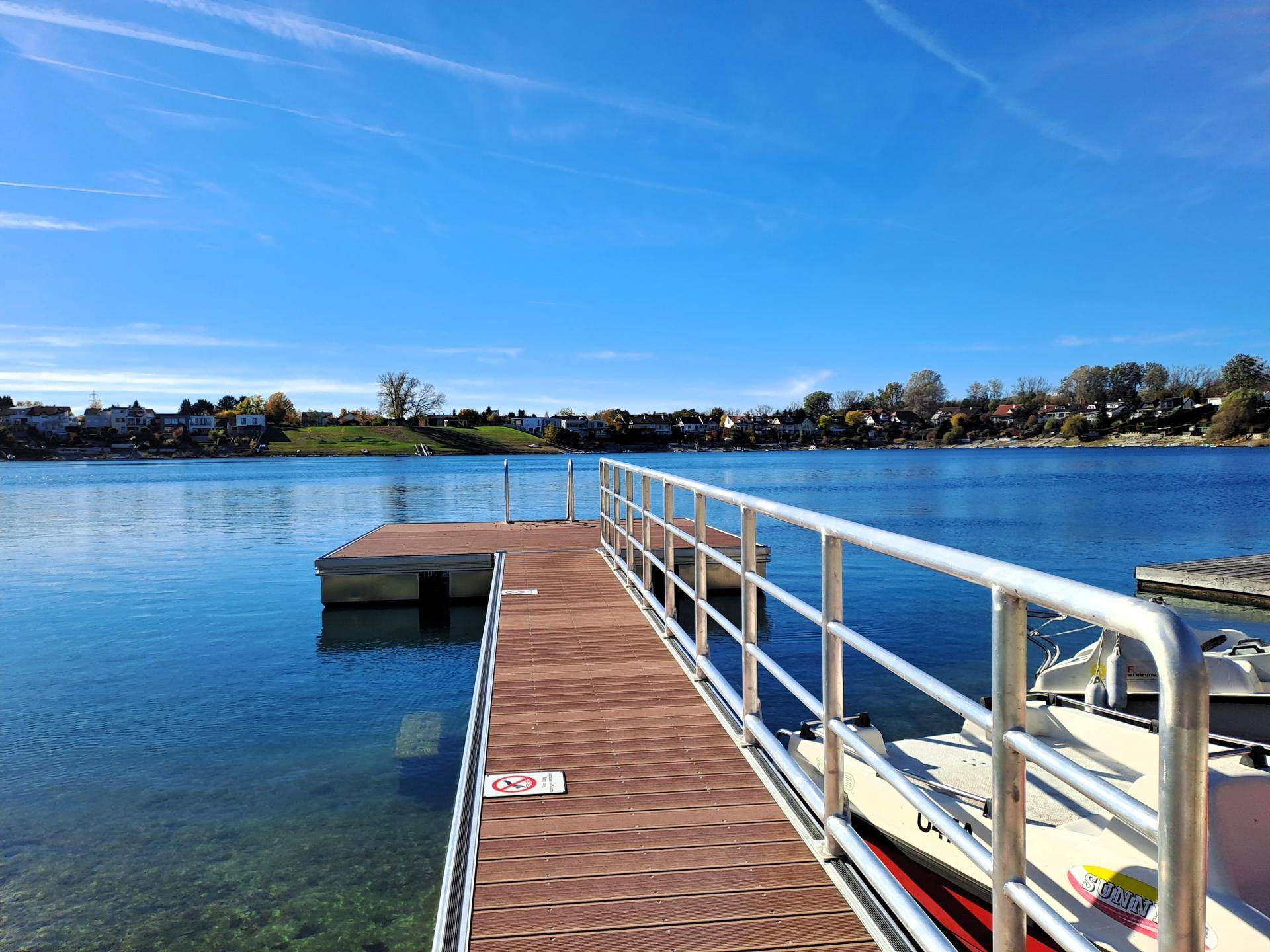Holzsteg am Seeufer mit Metallgeländer und kleinen Booten im ruhigen, blauen Wasser.