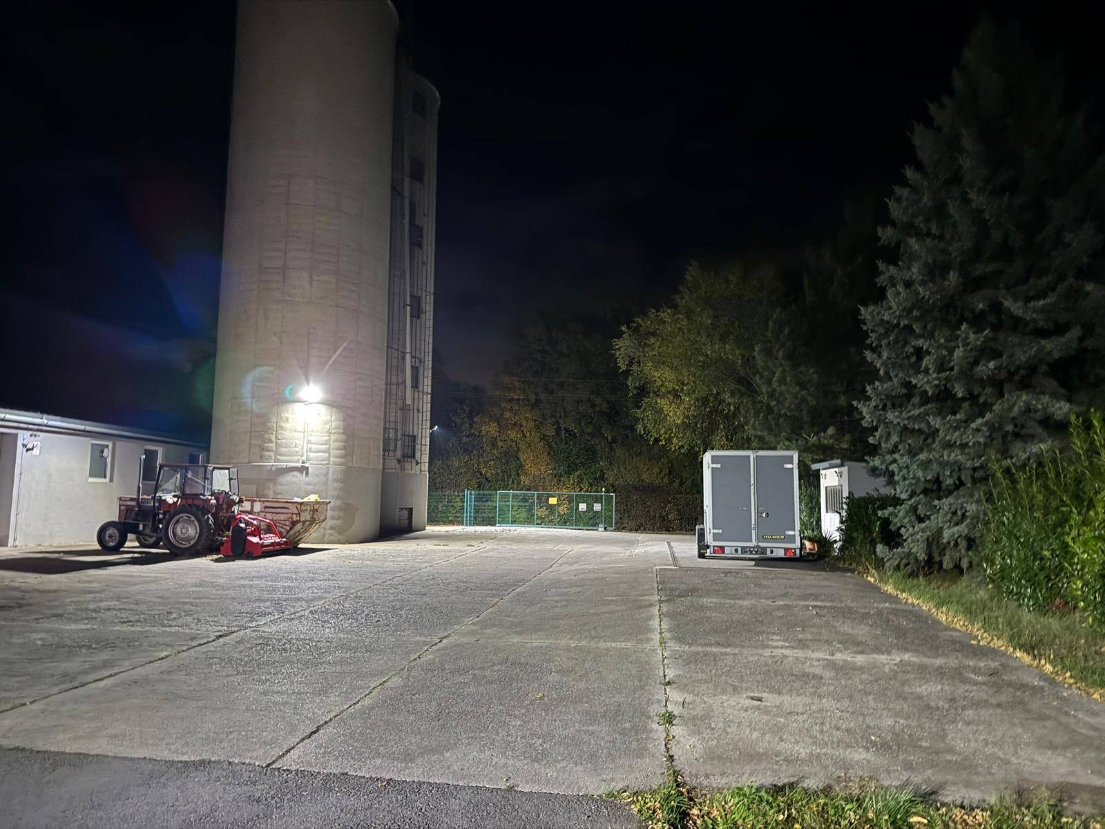 Nachtaufnahme des Geländes mit beleuchtetem Silo, Gebäuden und einem Traktor auf dem Vorplatz.
