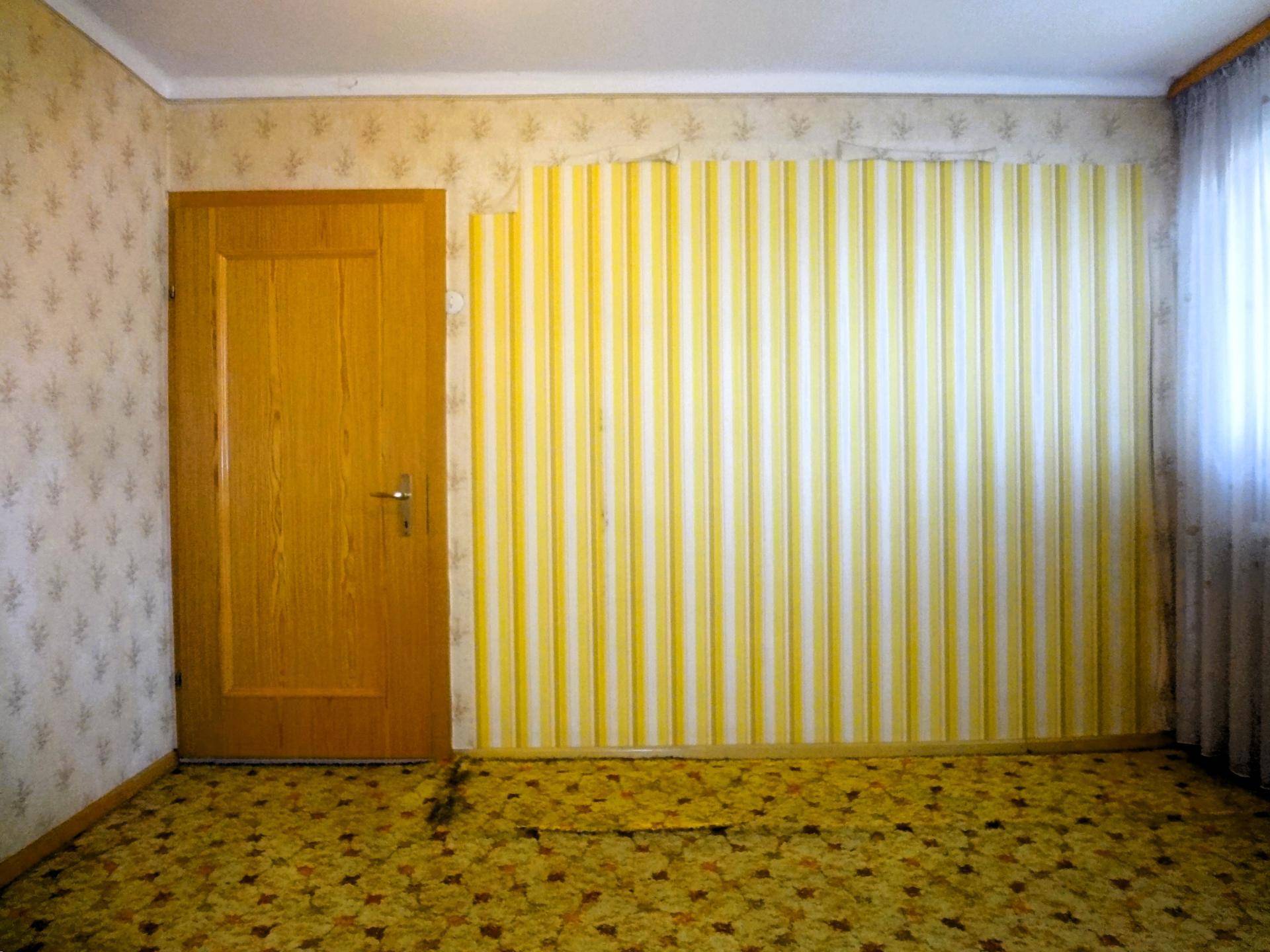 Schlafzimmer mit gemusterter Tapete, Teppichboden und Holztür.